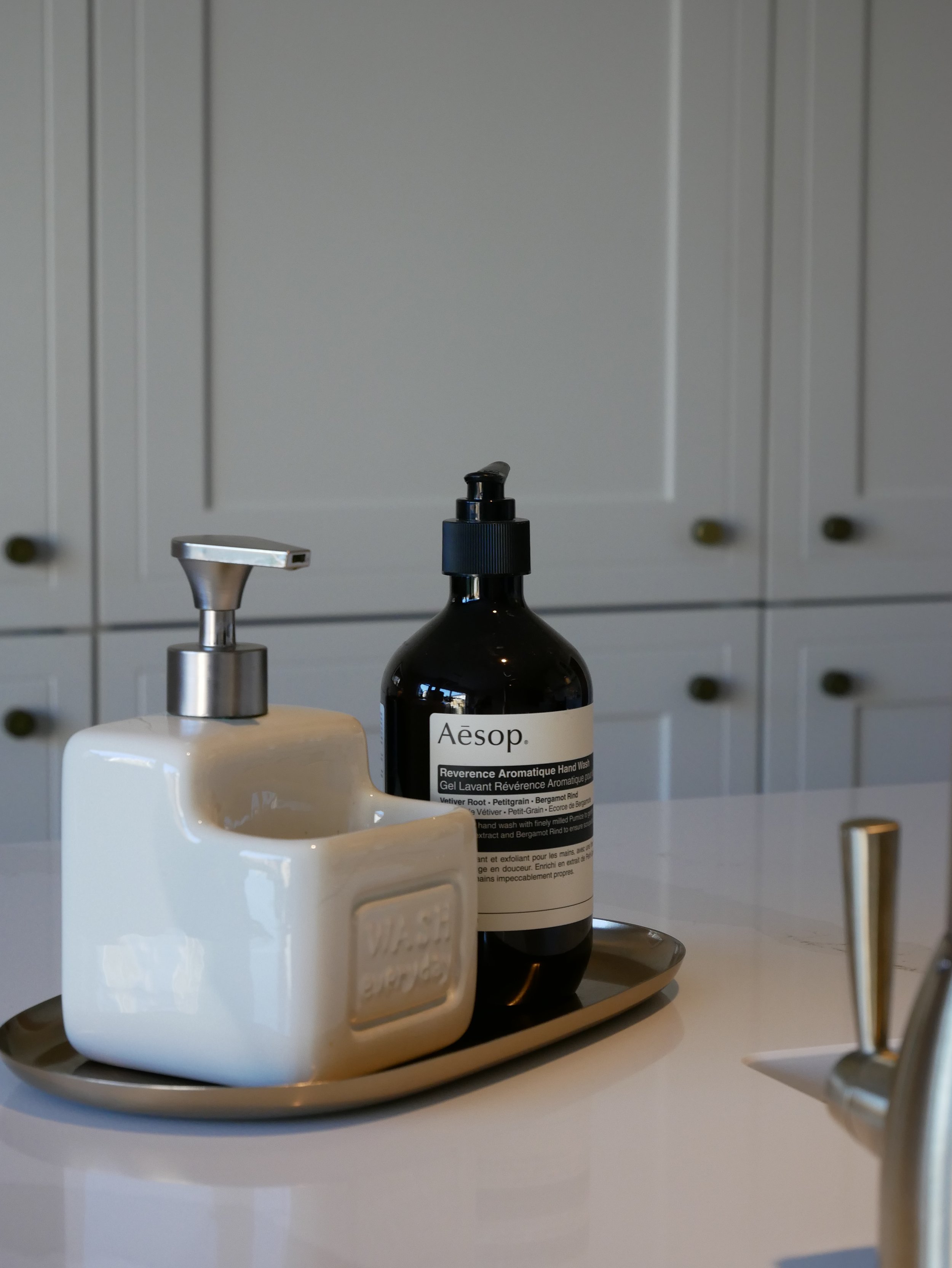 A tray with a white soap dispenser and a black bottle of Aesop hand wash on a white countertop, with gray cabinets in the background.