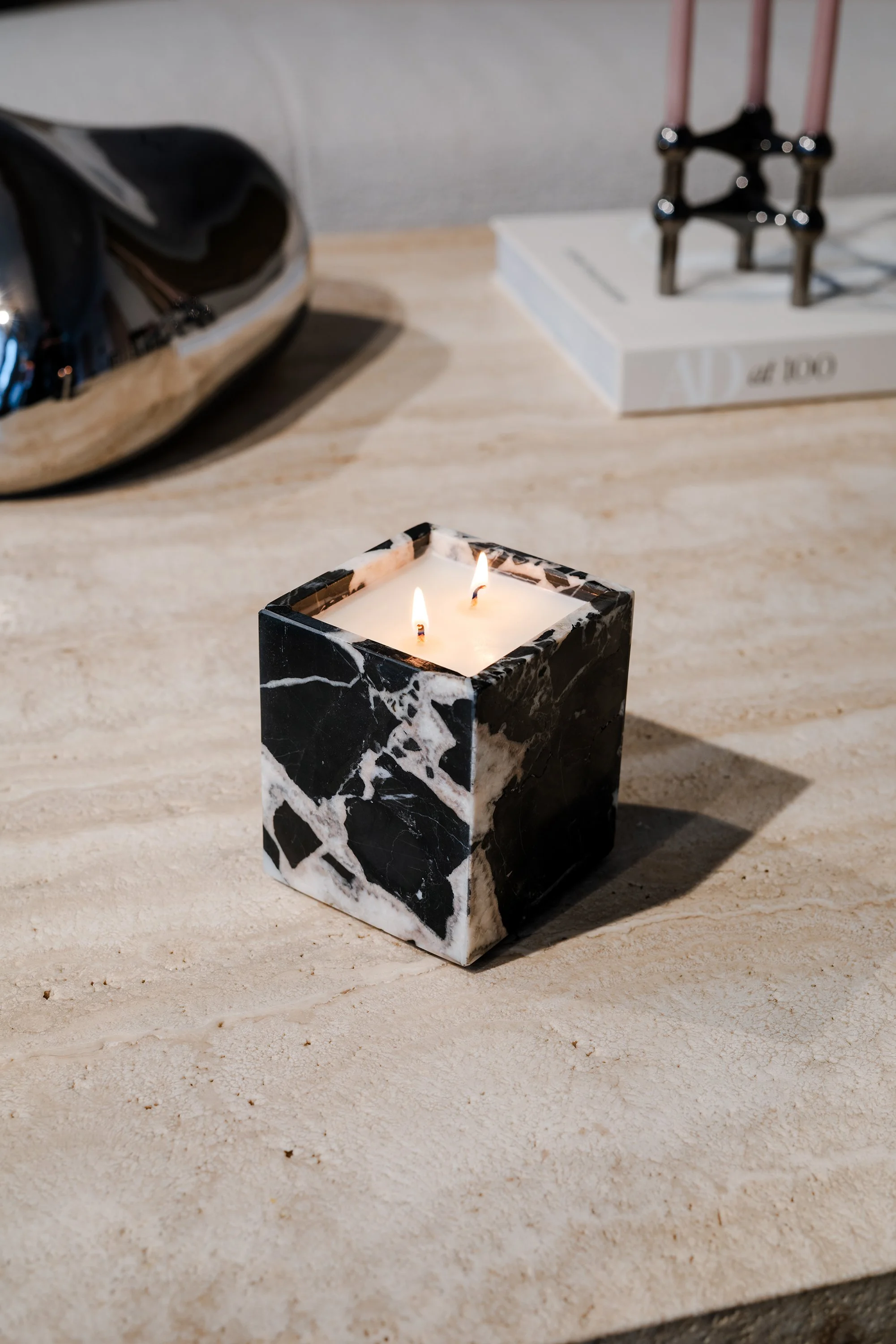 A black and white marble candle with two flames on a beige surface, with a book and abstract sculpture in the background.