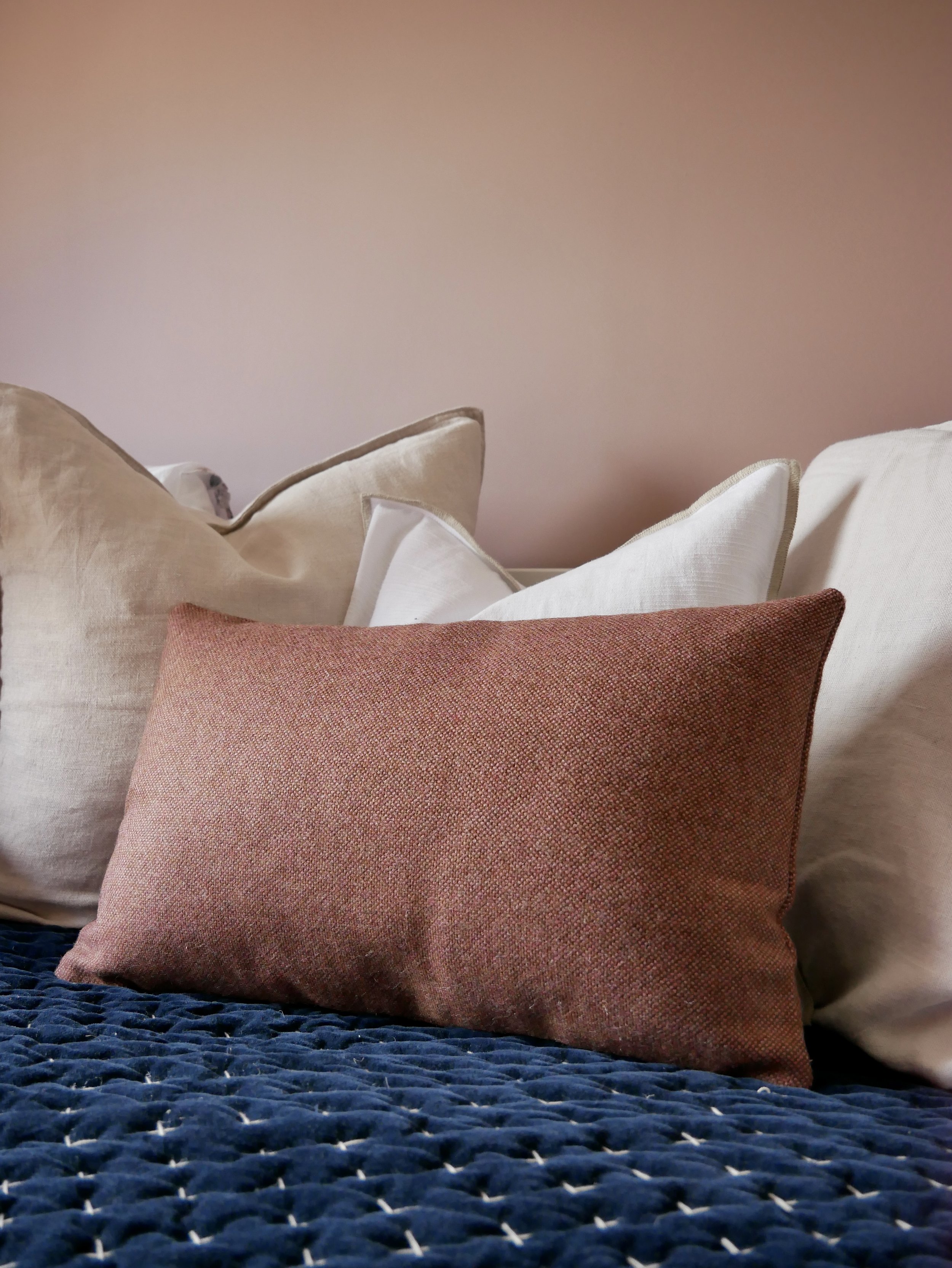 A collection of decorative pillows arranged on a bed with a textured blue blanket. The pillows are in various neutral shades, with the front pillow in a reddish-brown color. The background features a plain pink wall.