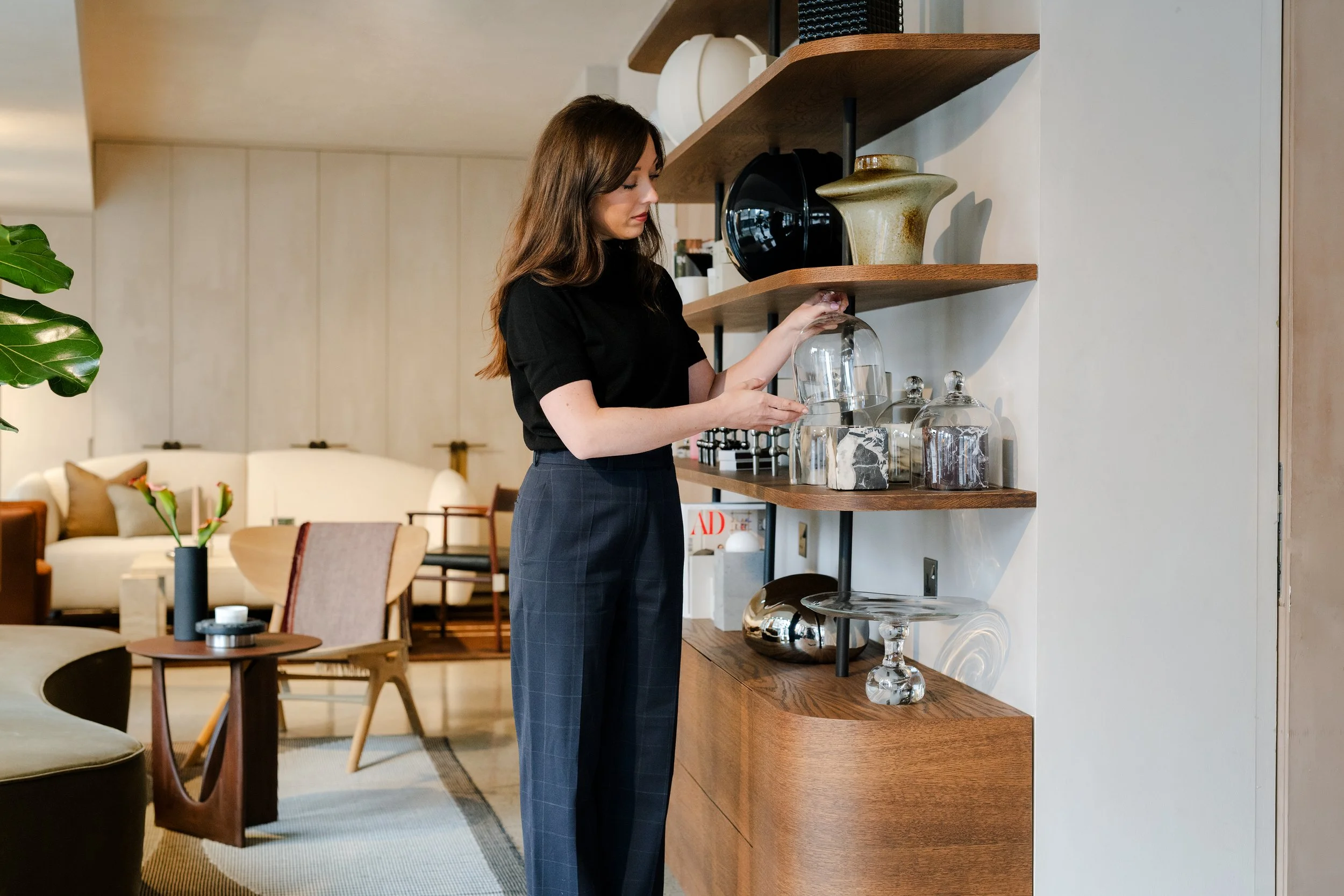 A woman with brown hair wearing a black shirt and blue plaid pants looks at a glass object on a wooden shelf in a modern living room or showroom.