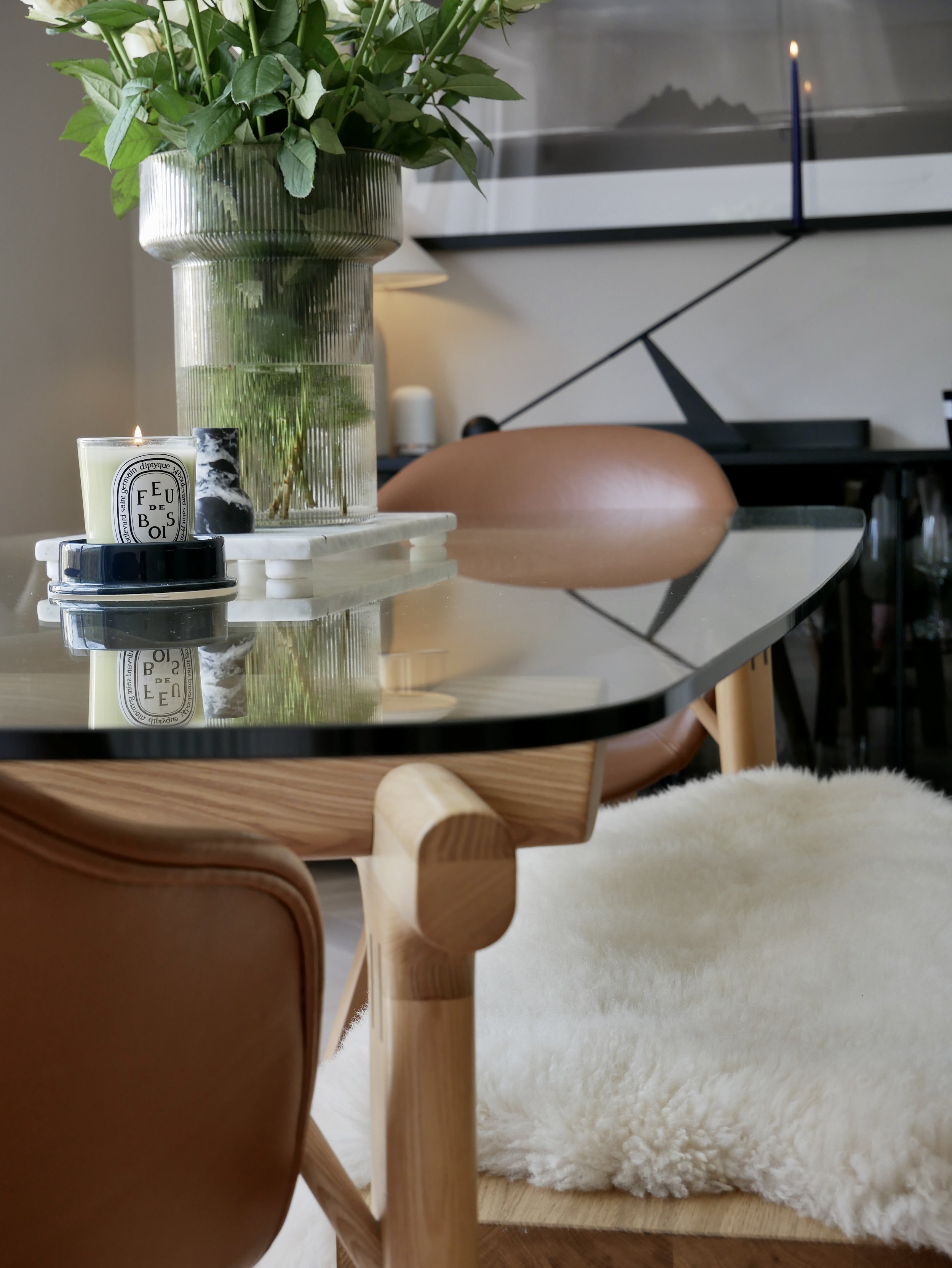 A glass-topped dining table with a large vase of green leaves, a lit candle, and a small decorative black and white stone, with three white marble coasters underneath. In the background, there is a brown leather and wood chair, a cream-colored fluffy