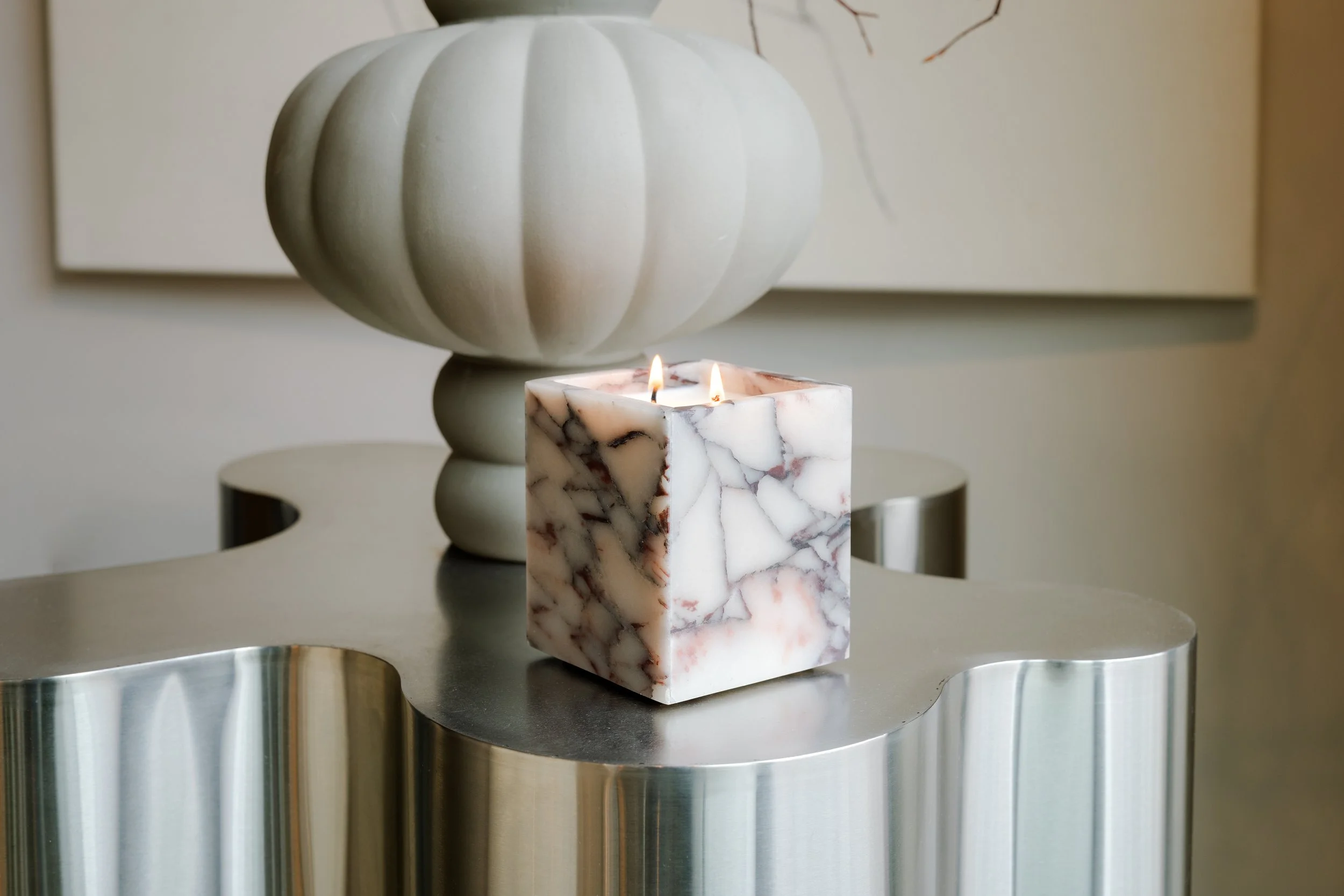 A white pumpkin-shaped vase, a lit marble-patterned candle, and a reflective metal surface.
