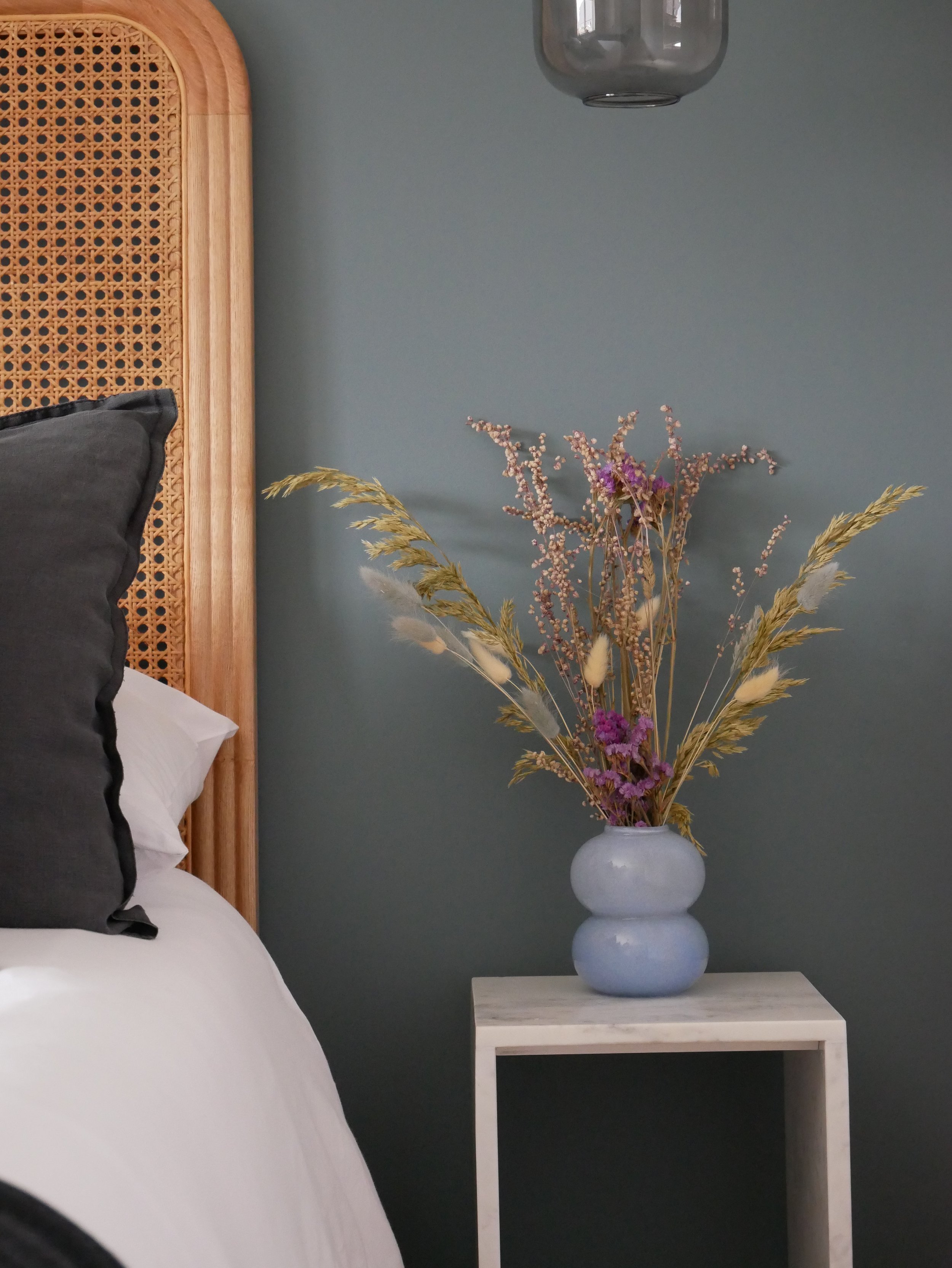 A white bedside table with a light gray vase holding dried flowers including purple, beige, and light pink blooms, against a blue-green wall. Part of a bed with a white sheet and black pillow is visible on the left.