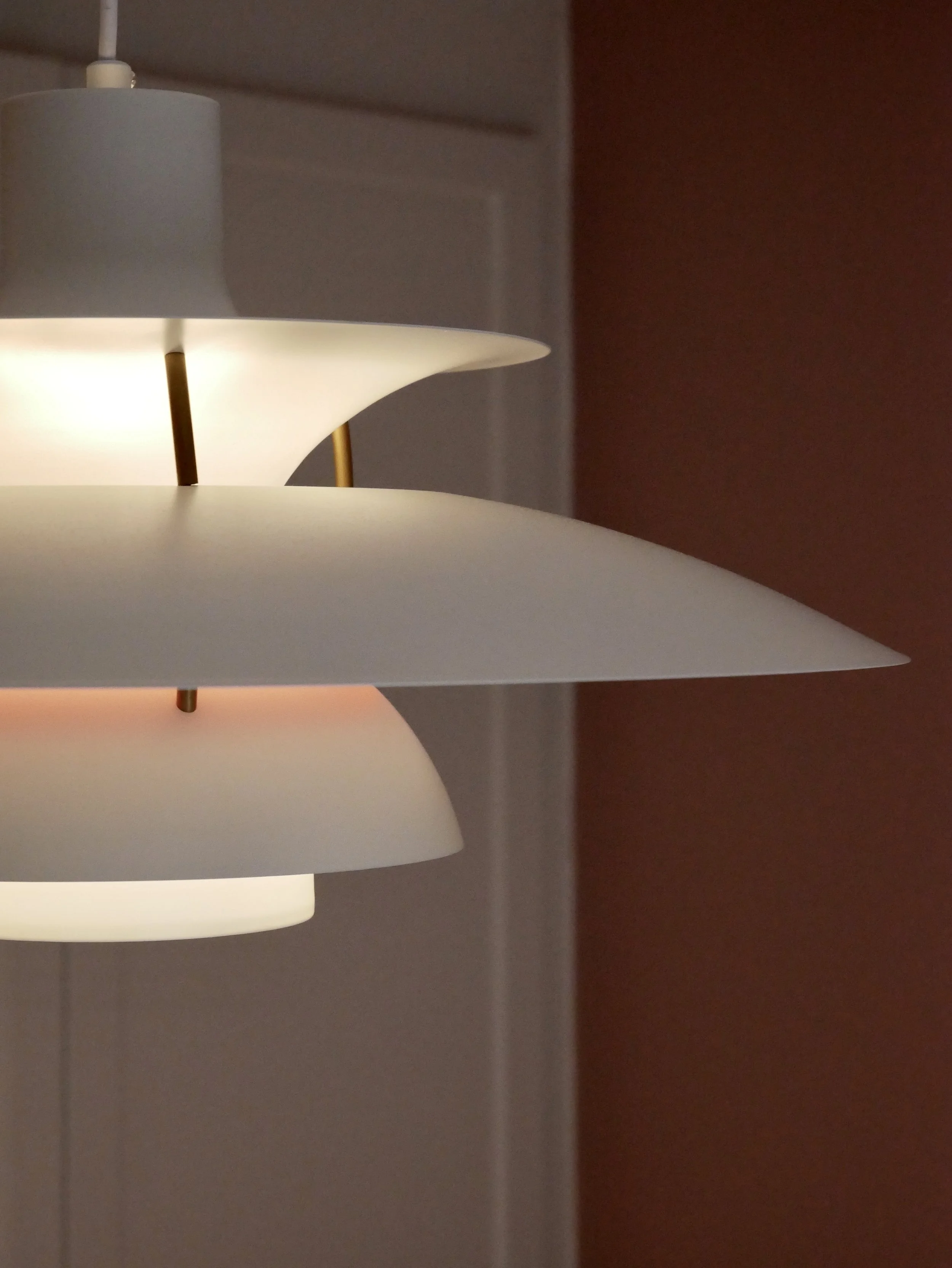 A modern, multi-tiered white and gold table lamp from Louis Poulsen with round, flat shades illuminated in a softly diffused way, standing against a wall with a dark red section and a door frame visible in the background.