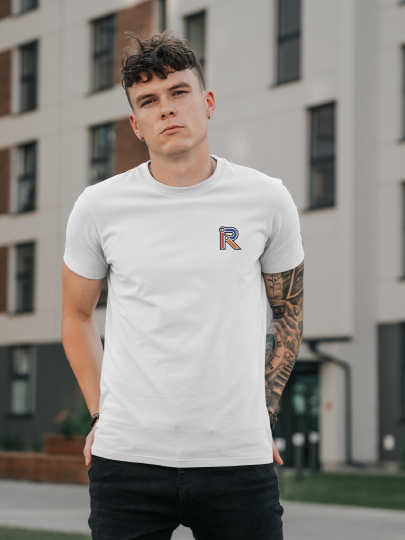 Young man with curly hair, wearing a white t-shirt with a small logo on the chest, standing outdoors in front of modern apartment buildings.