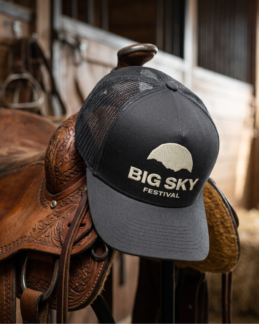 A black and gray trucker cap with the logo and text for Big Sky Festival, draped over a wooden saddle on a rustic barn wall.