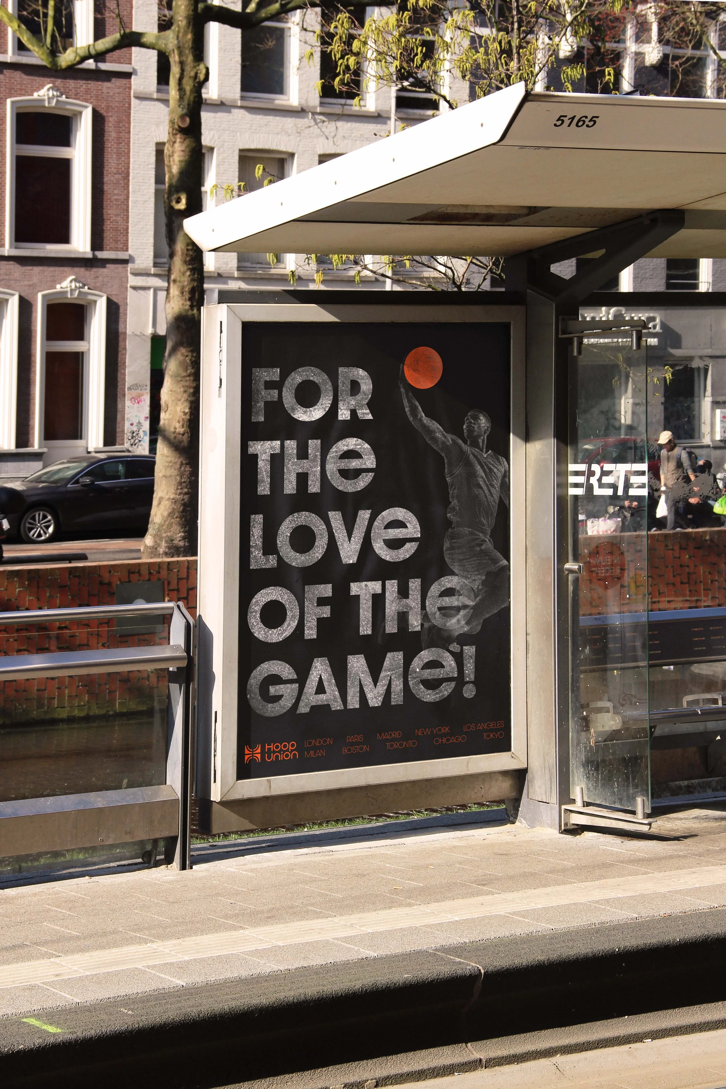 Bus stop shelter with a poster that reads 'FOR THE LOVE OF THE GAME!' featuring a basketball player silhouette holding a basketball, with city street and buildings in background.