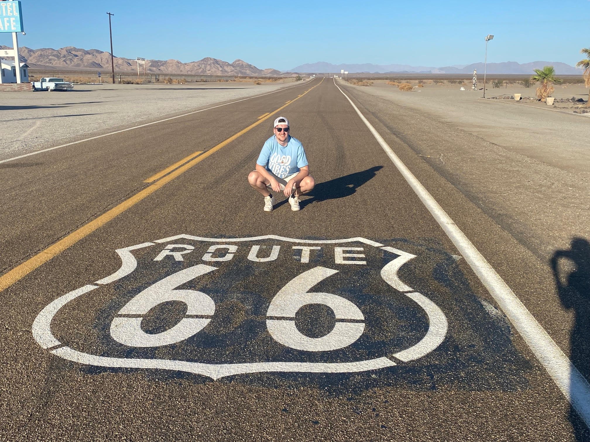 Person squatting on Route 66 road sign painted on the pavement of a desert highway with mountains in the background.