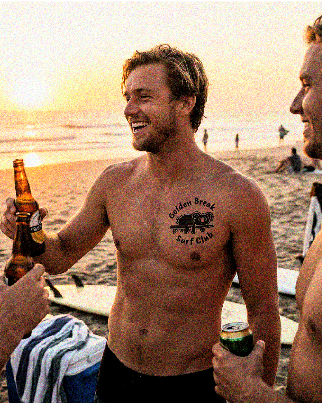 A group of men, shirtless, enjoying drinks and talking on the beach at sunset. One man has a tattoo that reads 'Golden Break Surf Club' with a koala bear in the middle.