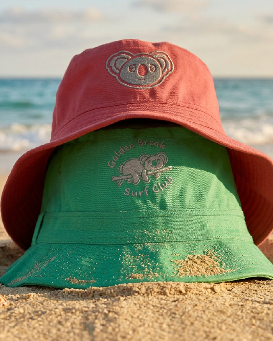 A pink wide-brimmed hat and a green bucket hat with koala bear designs and text, lying on sandy beach at the ocean.