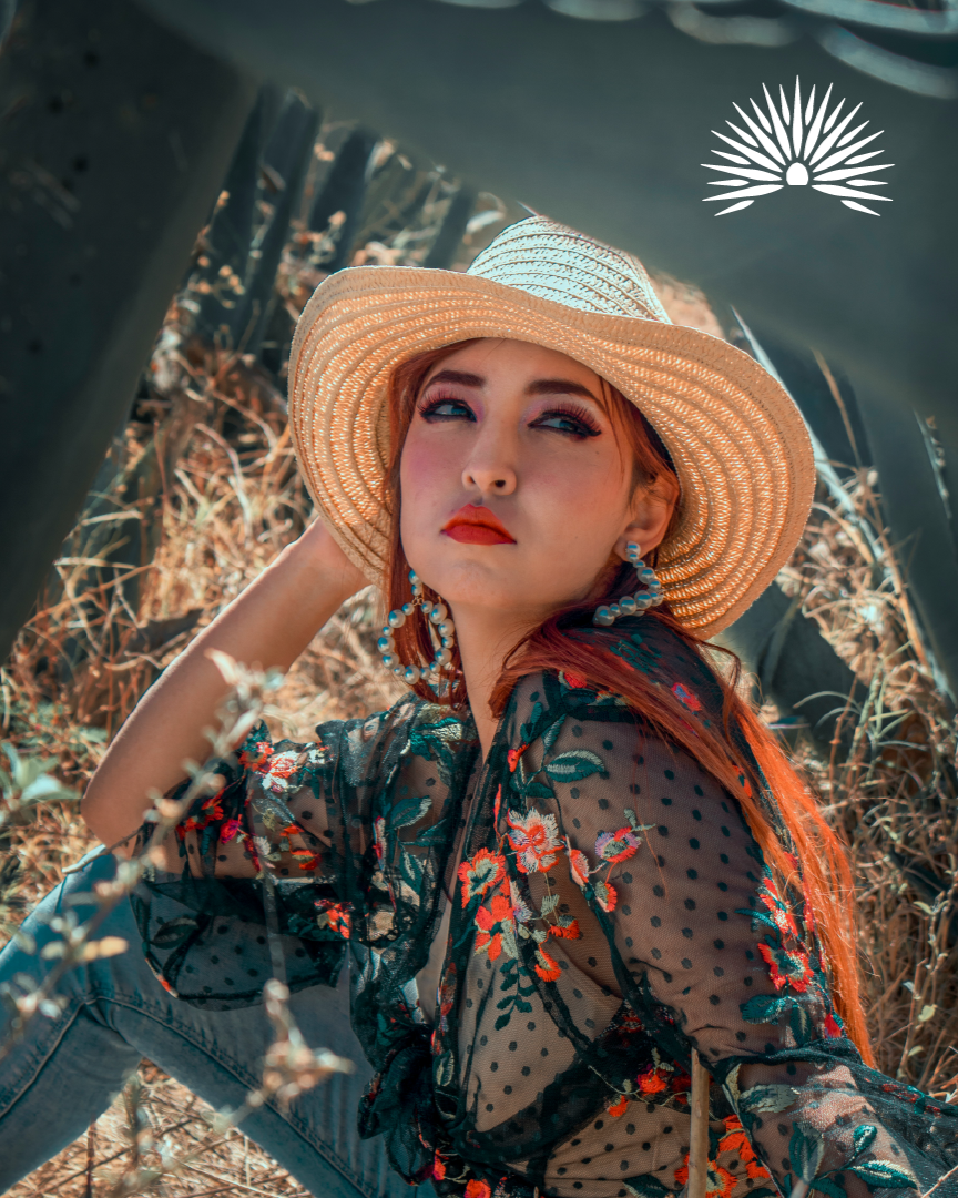 Young woman wearing a wide-brimmed straw hat, black sheer floral blouse, and earrings, sitting outdoors among tall grass and industrial structures.