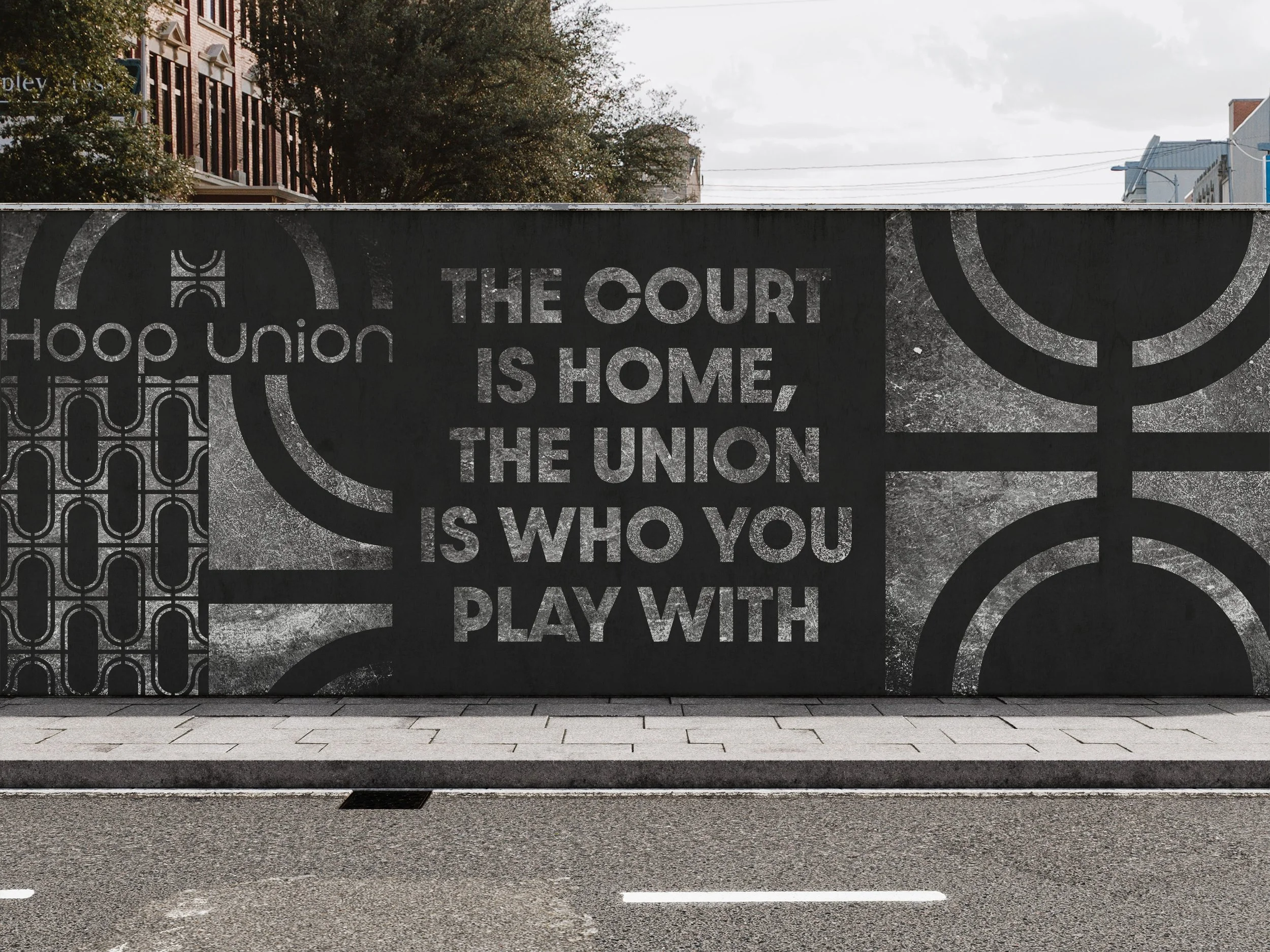 A mural with text that reads, "The court is home, the union is who you play with," featuring geometric and symbolic designs in black and white, located on a city street with buildings and trees in the background.