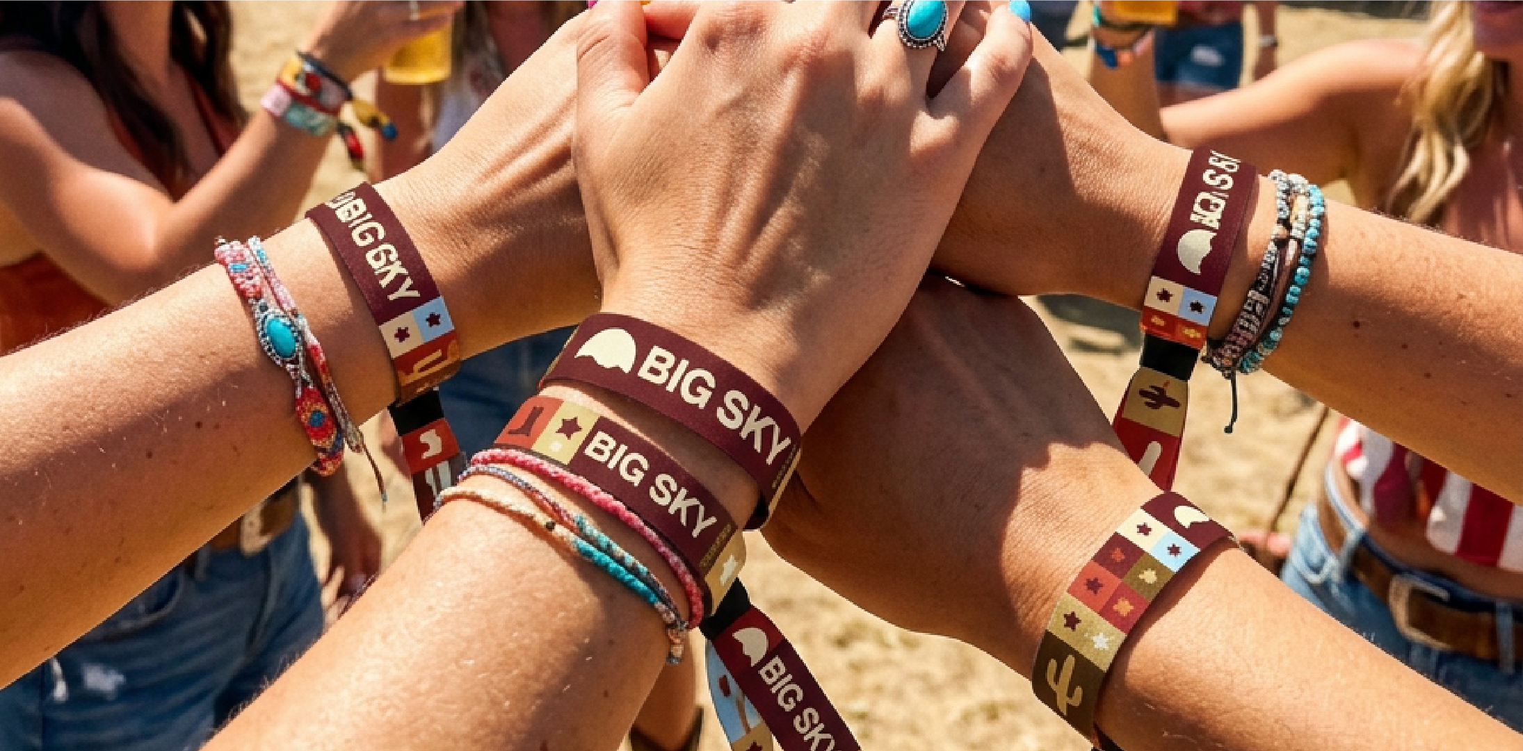 Group of people at an outdoor event placing their hands together in a stack, wearing festival wristbands and bracelets, with some holding drinks in the background.