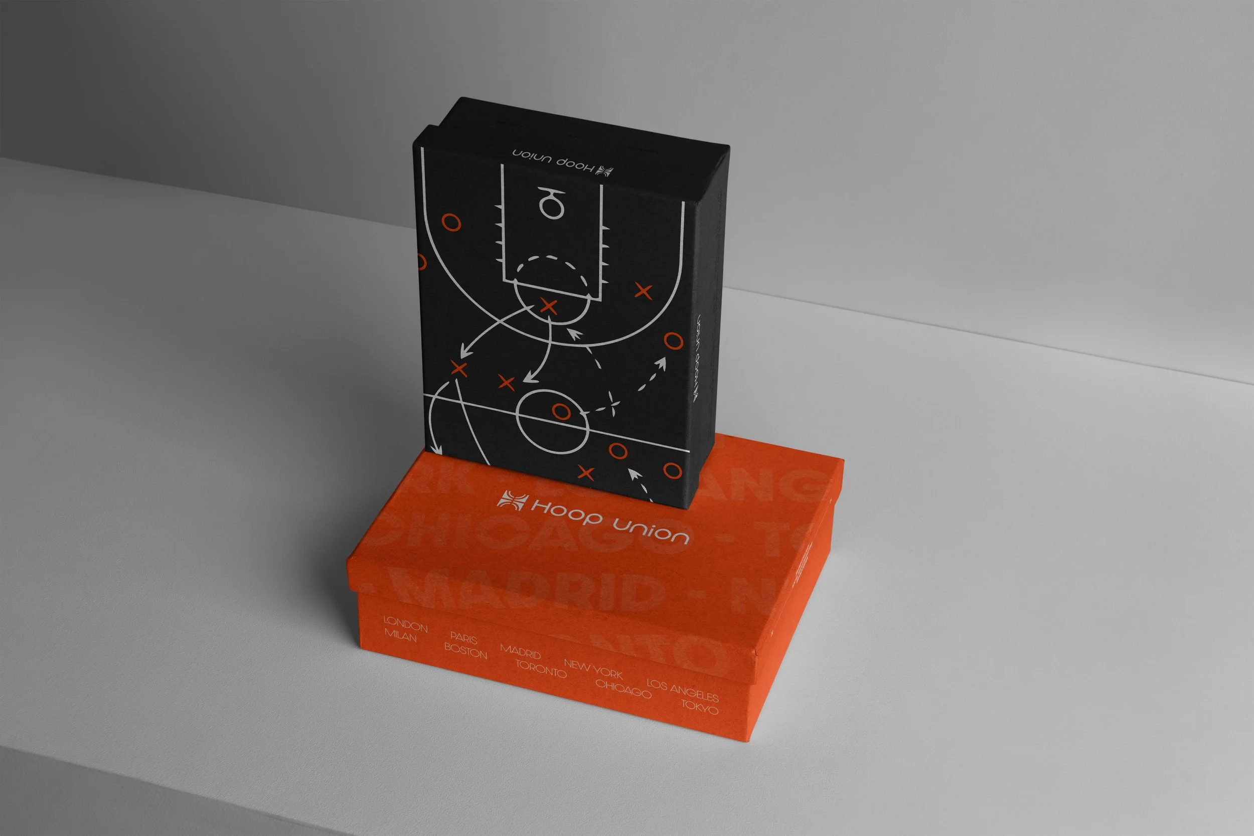 A black box with a sports strategy diagram and white and orange markings is displayed on top of an orange box with travel destination names printed in white. The black box features a basketball court diagram with arrows and circles, and is positioned at a slight angle against a white and gray background.