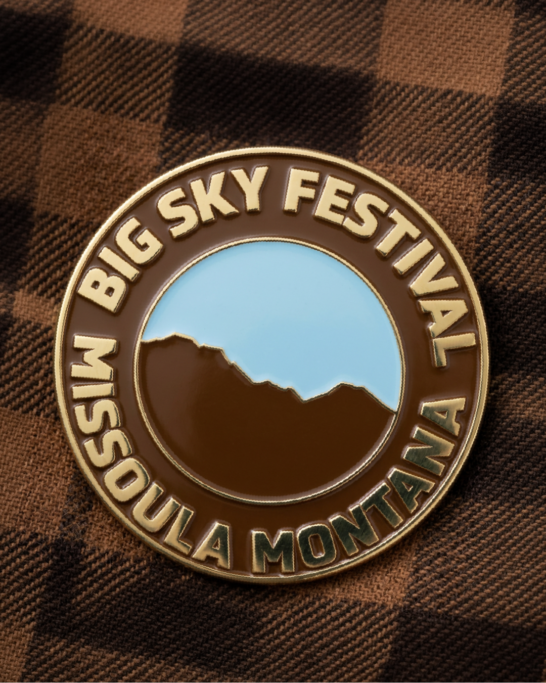 A commemorative pin for the Big Sky Festival, featuring the festivals logo and the festival's name encircling it, placed on a brown plaid fabric background.