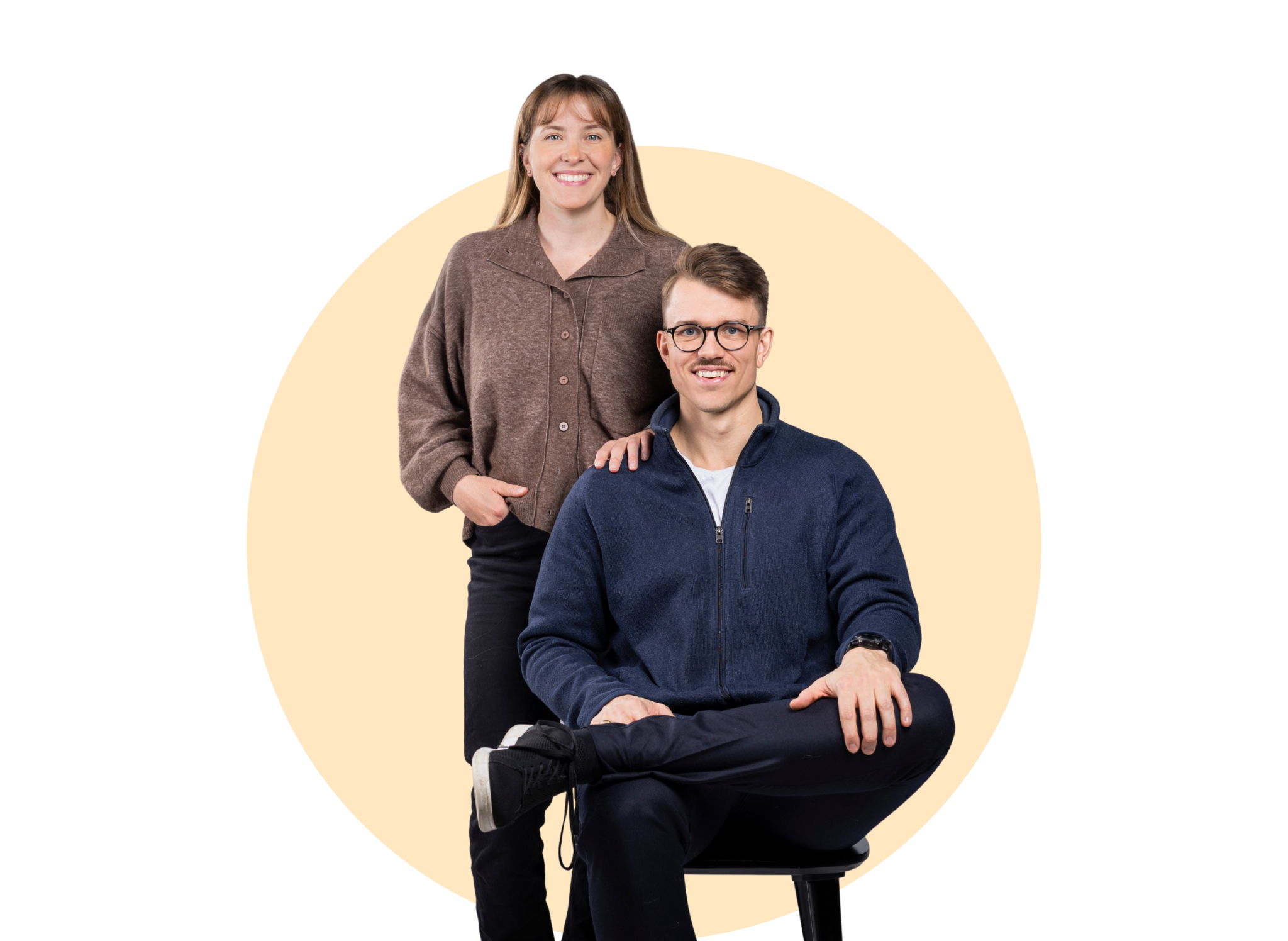A woman with long hair wearing a brown button-up shirt and a man with glasses, wearing a navy zip-up jacket, sitting on a stool, both smiling and facing the camera