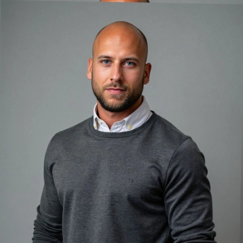 A man with a beard and short hair wearing a white collared shirt under a gray sweater, standing against a plain gray background.