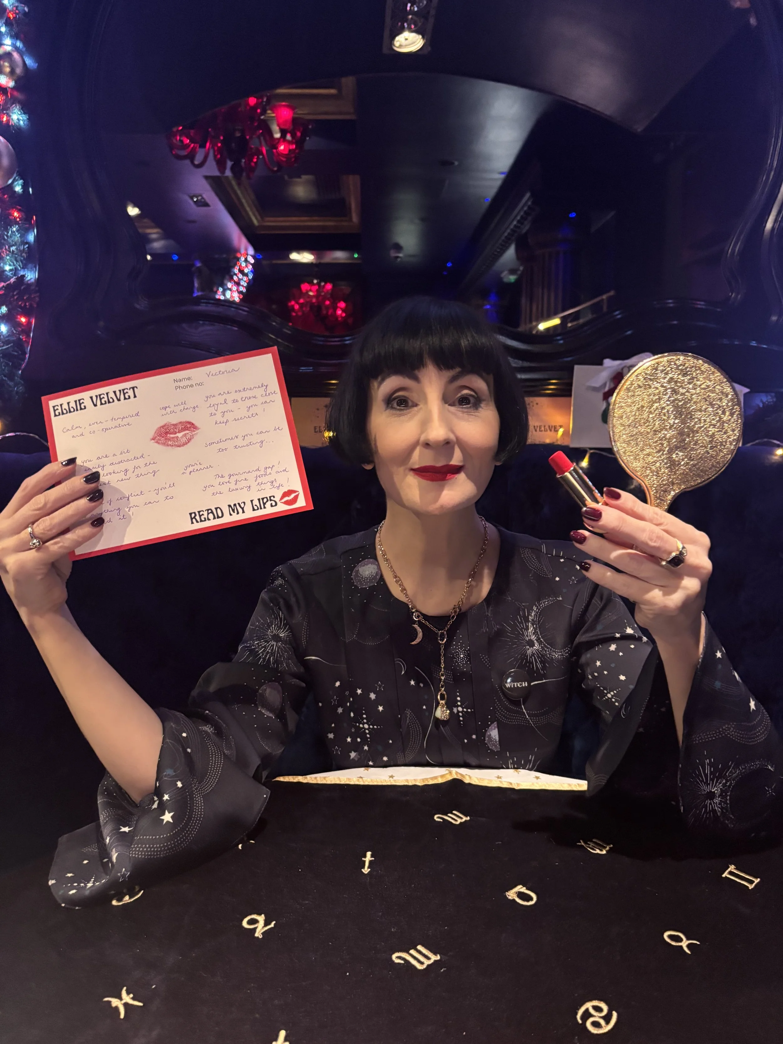 A woman with black hair and red lipstick holding a red lipstick tube and a gold mirror, smiling at the camera. She is sitting at a table with a black cloth decorated with gold letters. The background includes dark walls and red chandeliers, indicating an elegant setting that appears to be a bar or lounge.