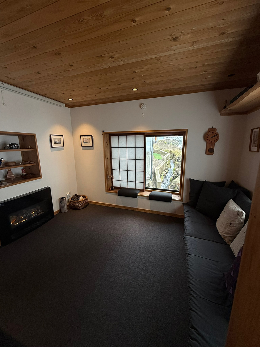 A cozy living room with a wooden ceiling, a window with a view outside, a black sofa with pillows, wall art, a wall clock, and a corner electric fireplace.