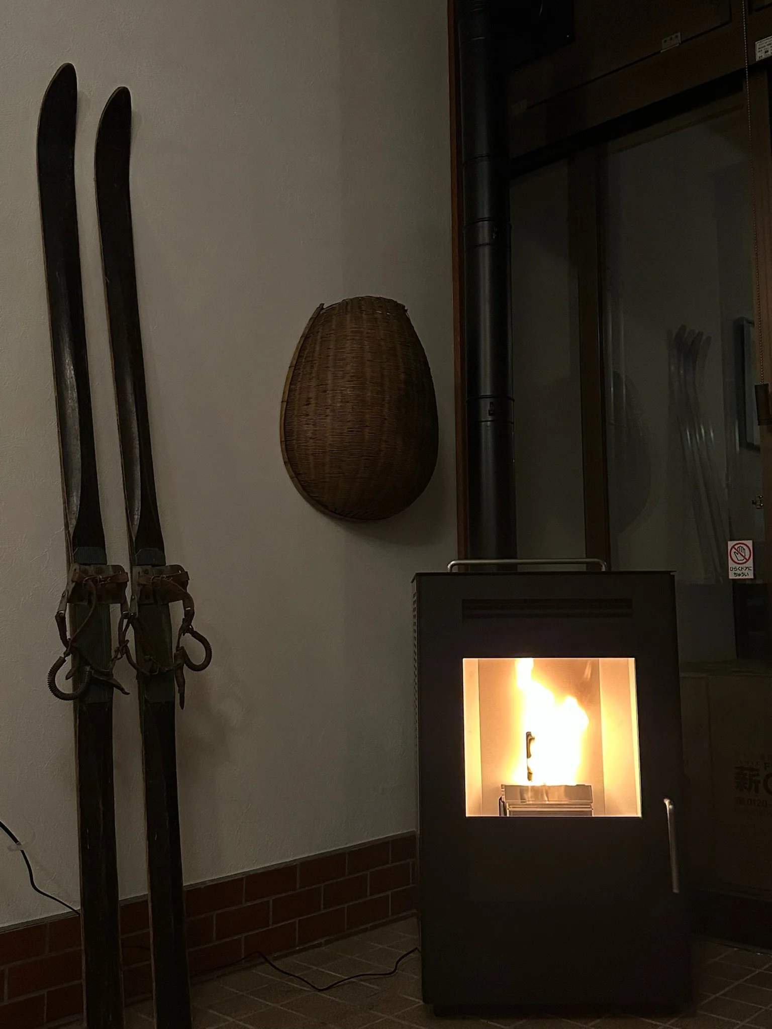 A set of vintage skis leaning against a wall next to a rattan wall basket, with a small black stove burning inside, emitting flames.