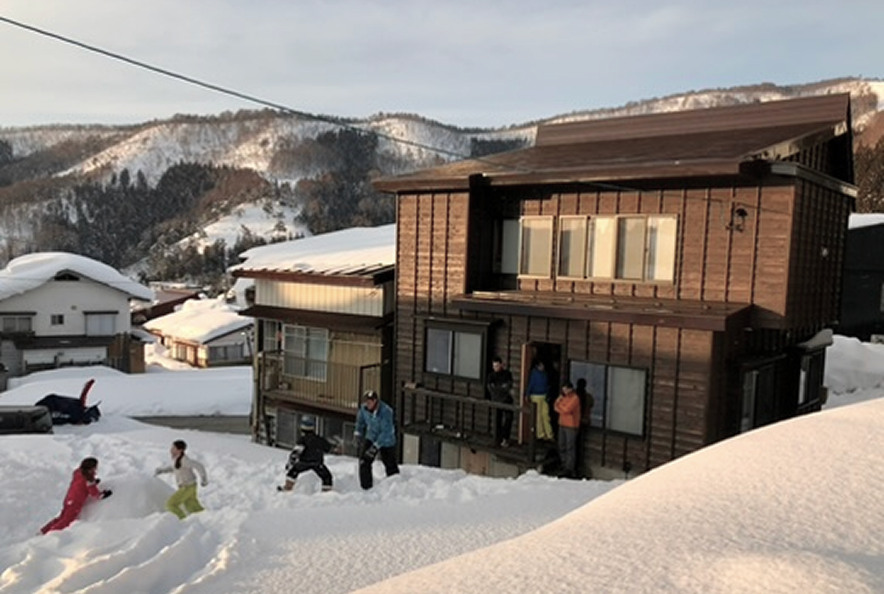 People playing in the snow outside wooden houses in a snowy mountain residential area.
