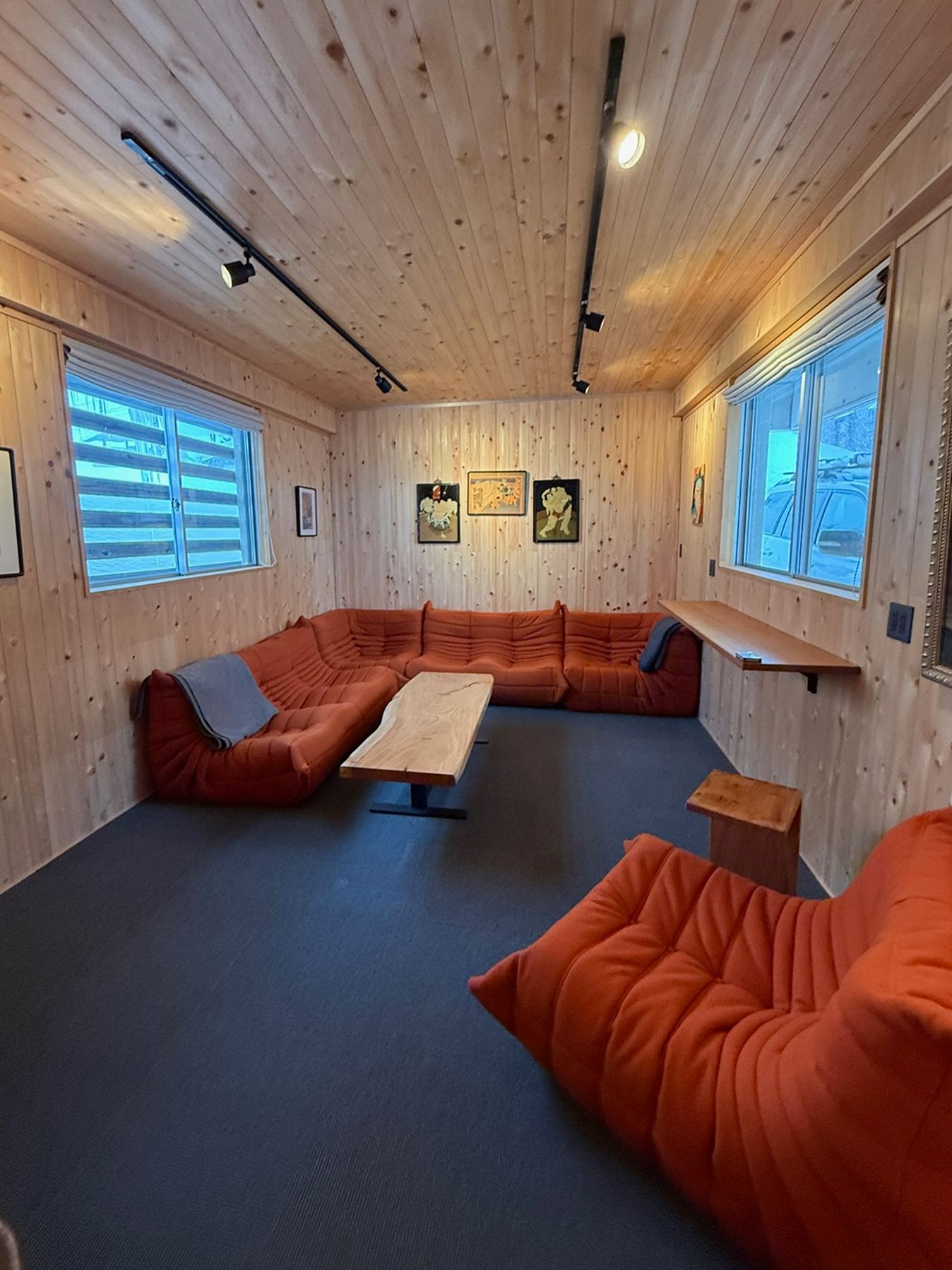 A cozy living room with wood-paneled walls and ceiling, featuring an L-shaped orange sectional sofa, a wooden coffee table, and artwork on the wall. Large windows with blinds let in natural light.