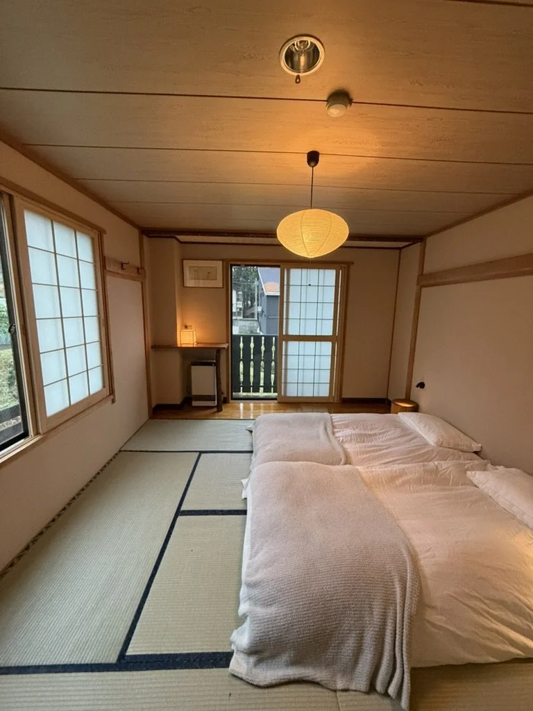 A cozy Japanese-style room with tatami mats, a futon bed, shoji paper sliding doors, a small desk with a lamp, and a balcony.