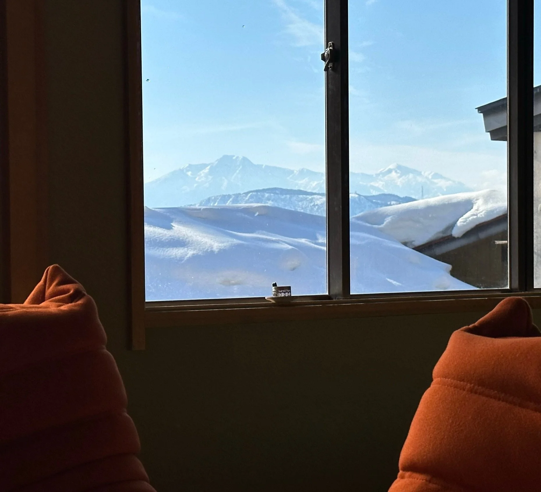 A view through a window showing snow-covered mountains and landscape, with brown pillows or seats visible in the foreground inside a cozy room.