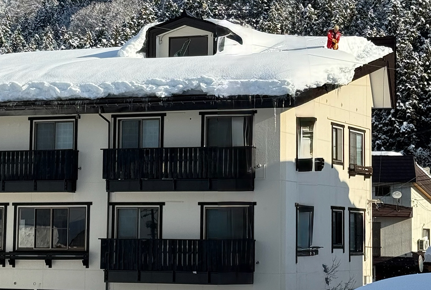 A multi-story white apartment building covered in snow with black balconies and windows, with snow piled on the roof and a person in winter gear standing on the roof.