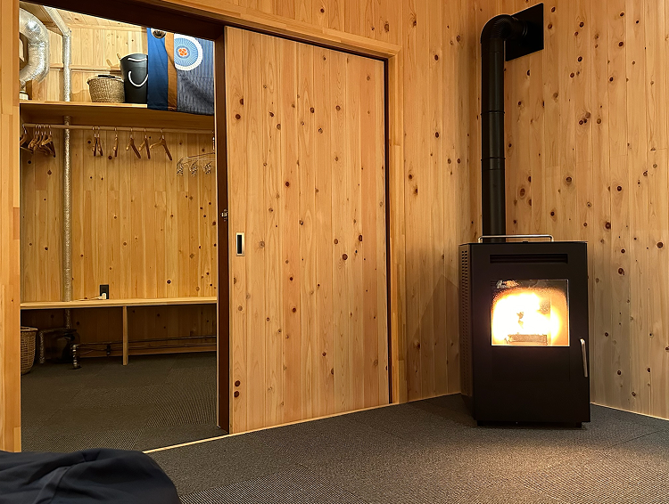 A room with wood-paneled walls and a black wood stove with a lit fire, a black chimney pipe, and an open sliding door revealing a closet with shelves, baskets, and hanging rods.