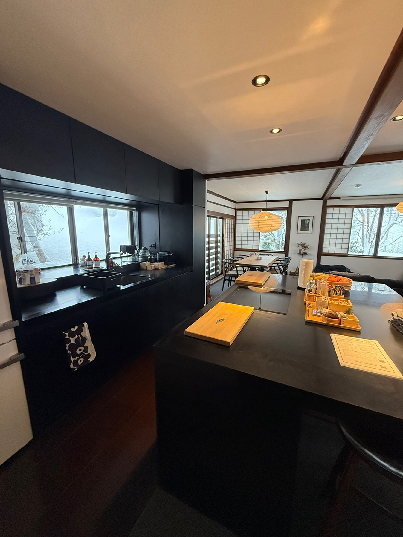 Interior of a modern kitchen with black cabinets, a large island with wooden cutting boards, and a dining area with a paper lantern ceiling light and shoji-style windows.