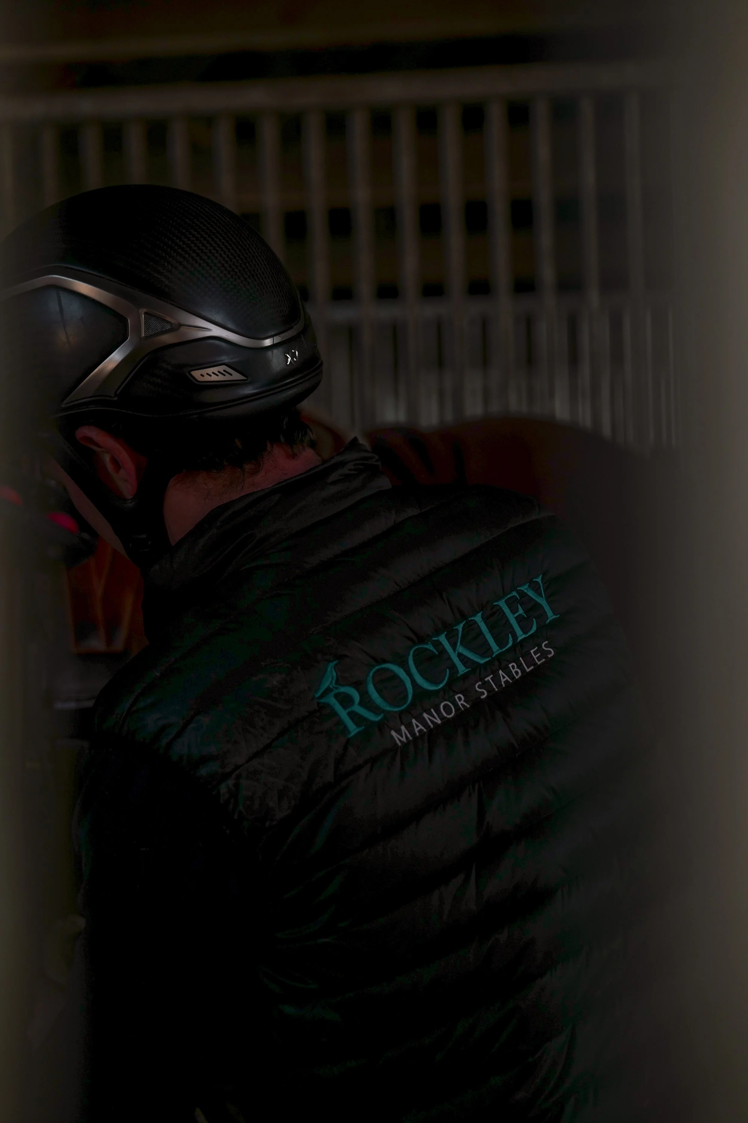 Person wearing a black motorcycle helmet and a black jacket with the logo 'Rockleys Manor Stables' on the back, standing indoors.