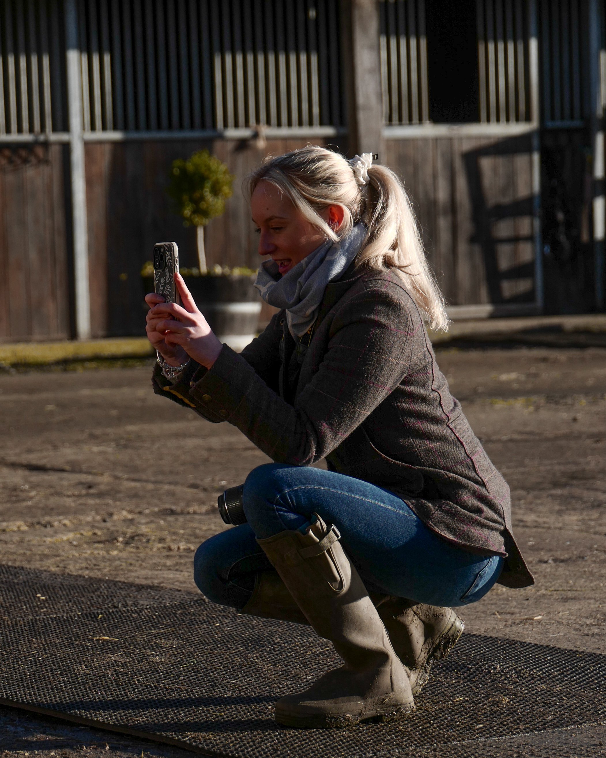 A woman squatting outdoors, taking a photo with her smartphone, wearing a dark jacket, jeans, and tall boots, with a potted plant and wooden fence in the background.