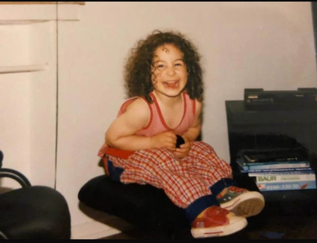 A young girl with curly hair smiling, sitting on a cushion with her knees up and feet on the floor, wearing a red plaid outfit and red sneakers, indoors.