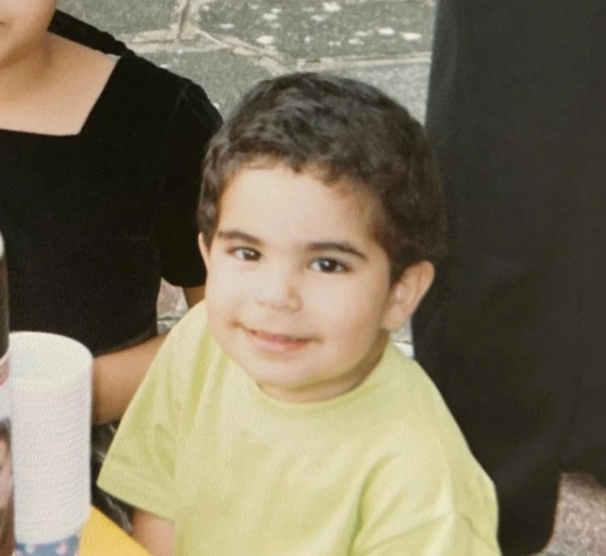 A young boy with dark hair smiling, wearing a yellow shirt, sitting at a table during a gathering or celebration.