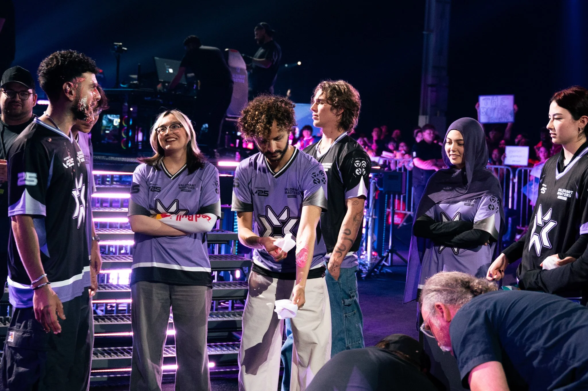 Group of esports team members in jerseys, some smiling, some serious, standing in a gaming arena with stage lights and equipment in the background.
