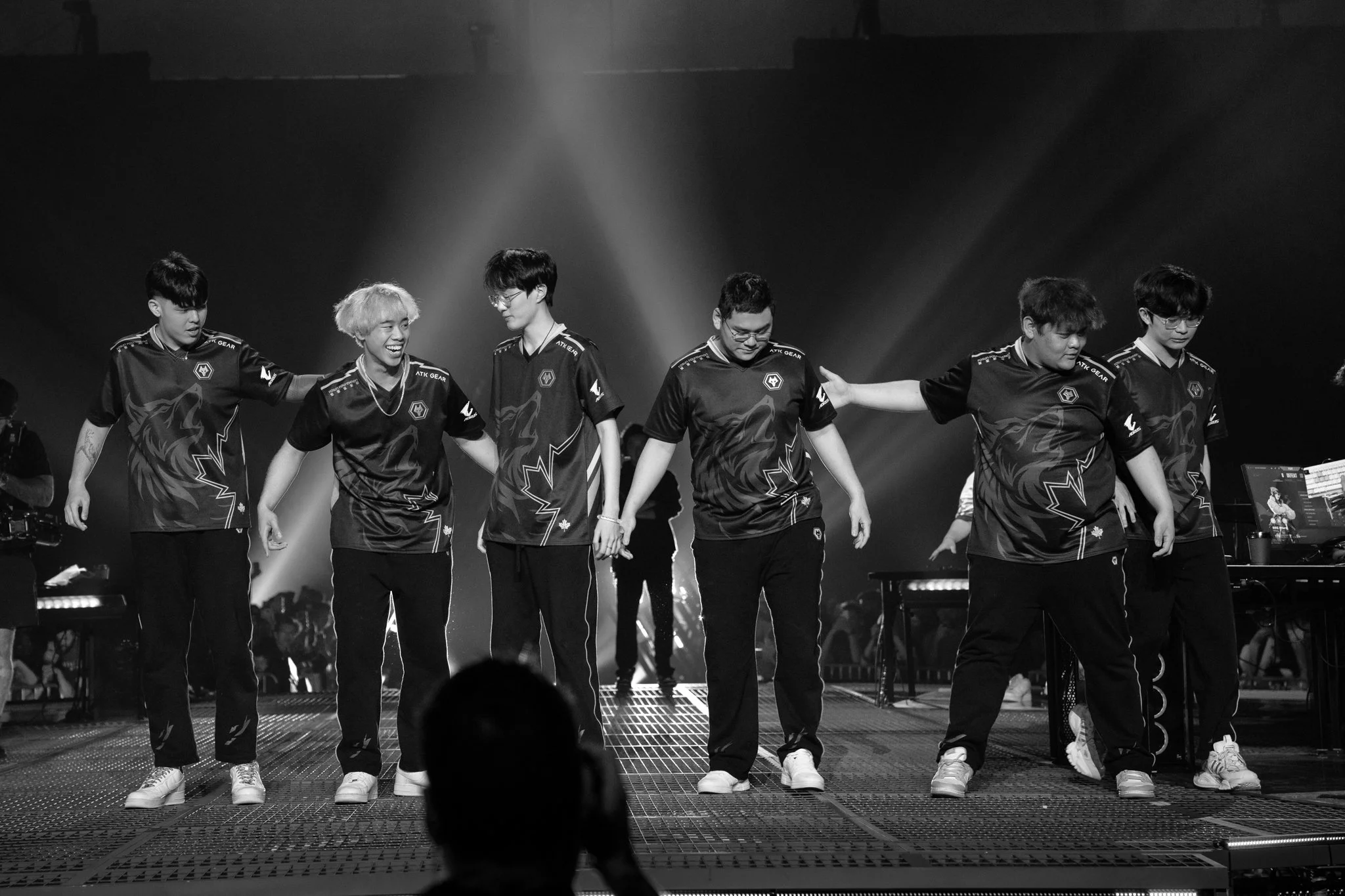 A group of six young men wearing matching esports team jerseys, holding hands and taking a bow on stage during a gaming event.