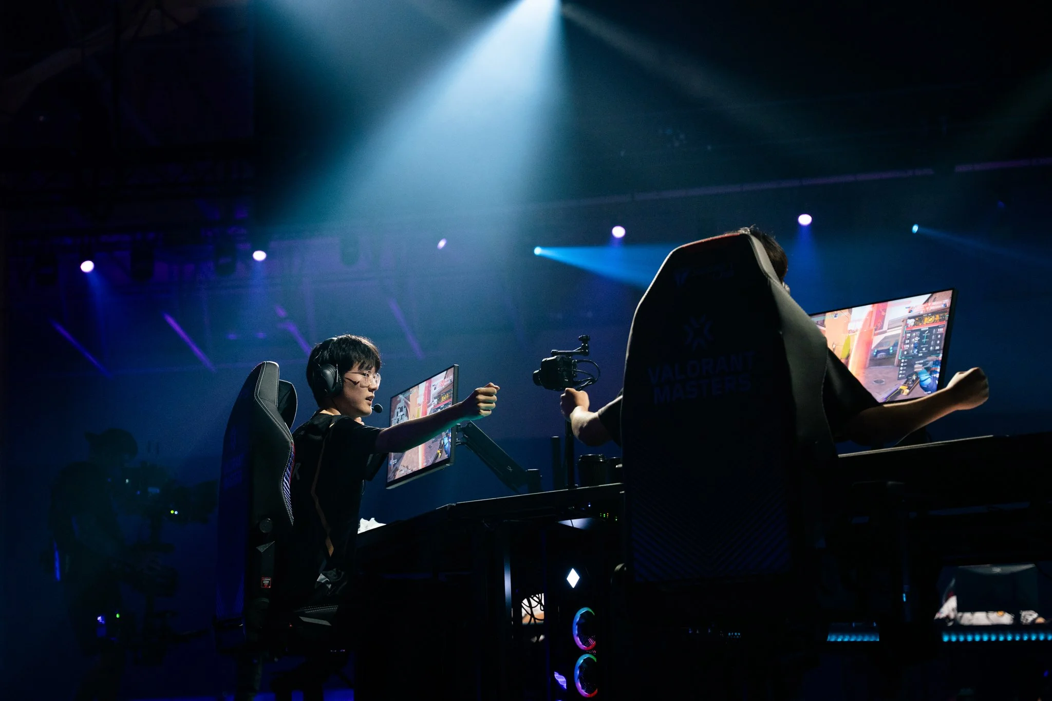 E-sports players at a gaming competition with illuminated monitors, gaming chairs, and stage lighting.