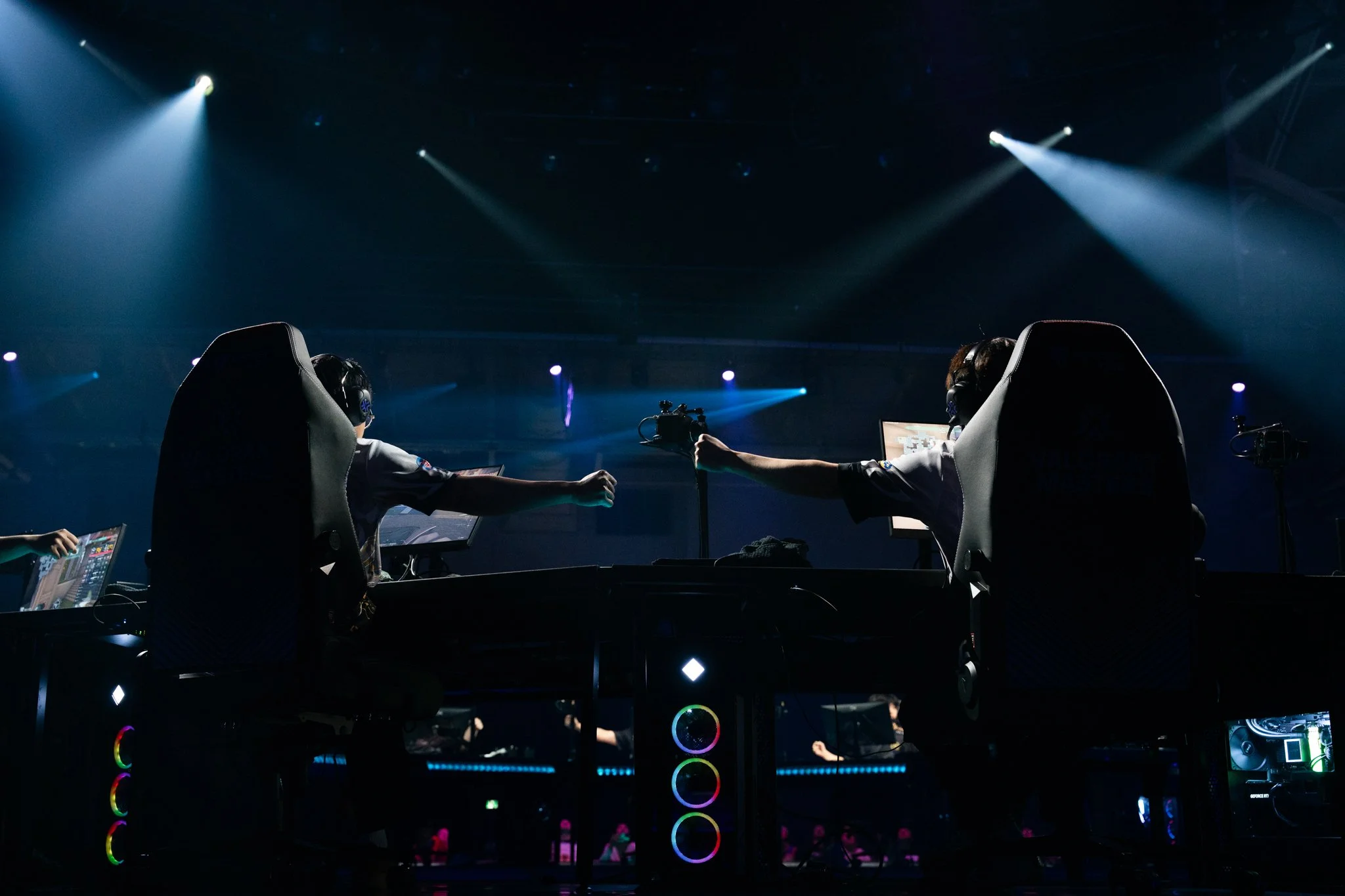 Two electronic esports players wearing headsets and gaming jerseys, reaching out to shake hands over gaming computers in a dark, illuminated arena with spotlights.