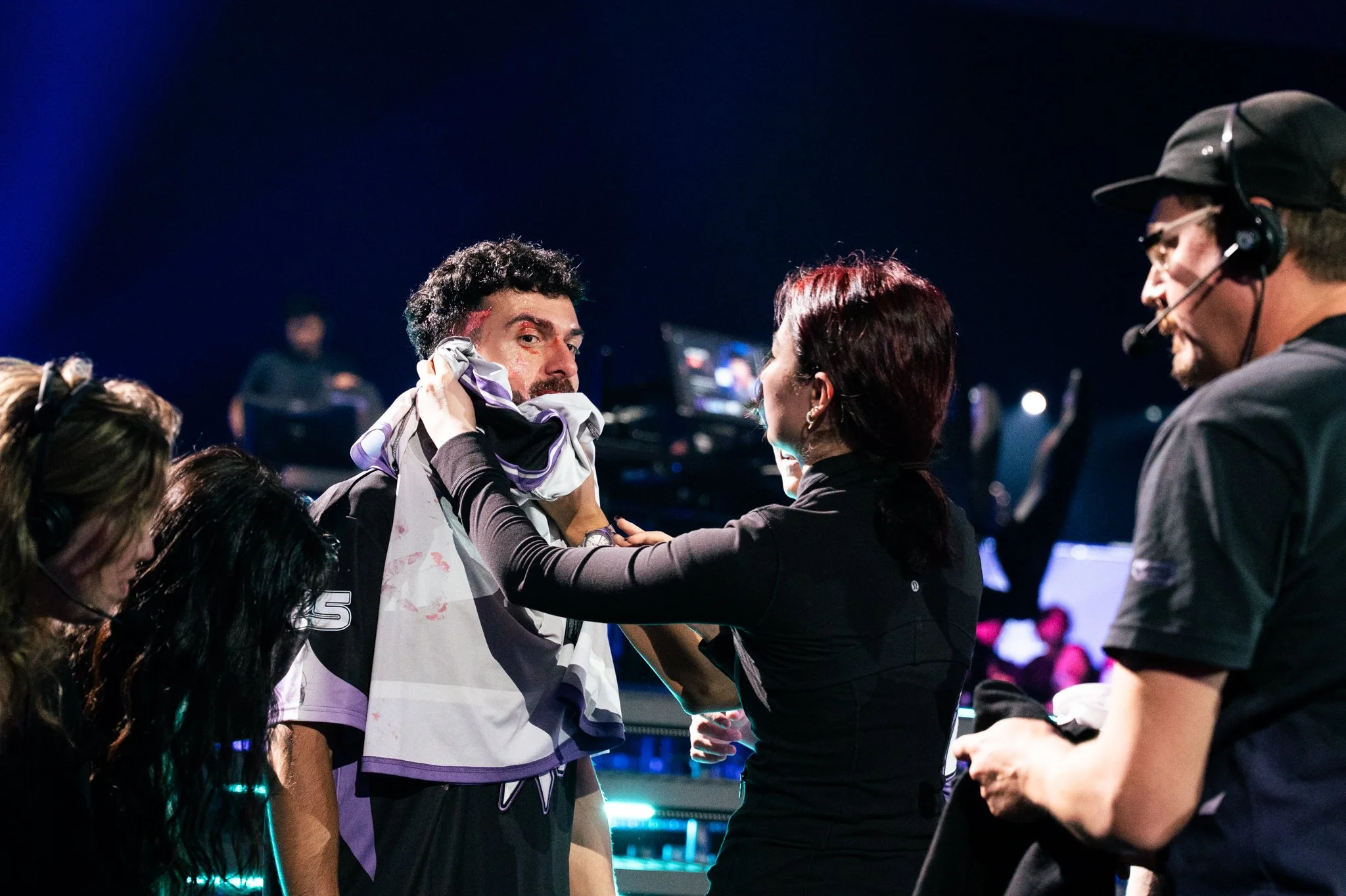 A person being decorated with a purple and white cloth by a woman backstage at an event, with crew members and equipment visible in the background.