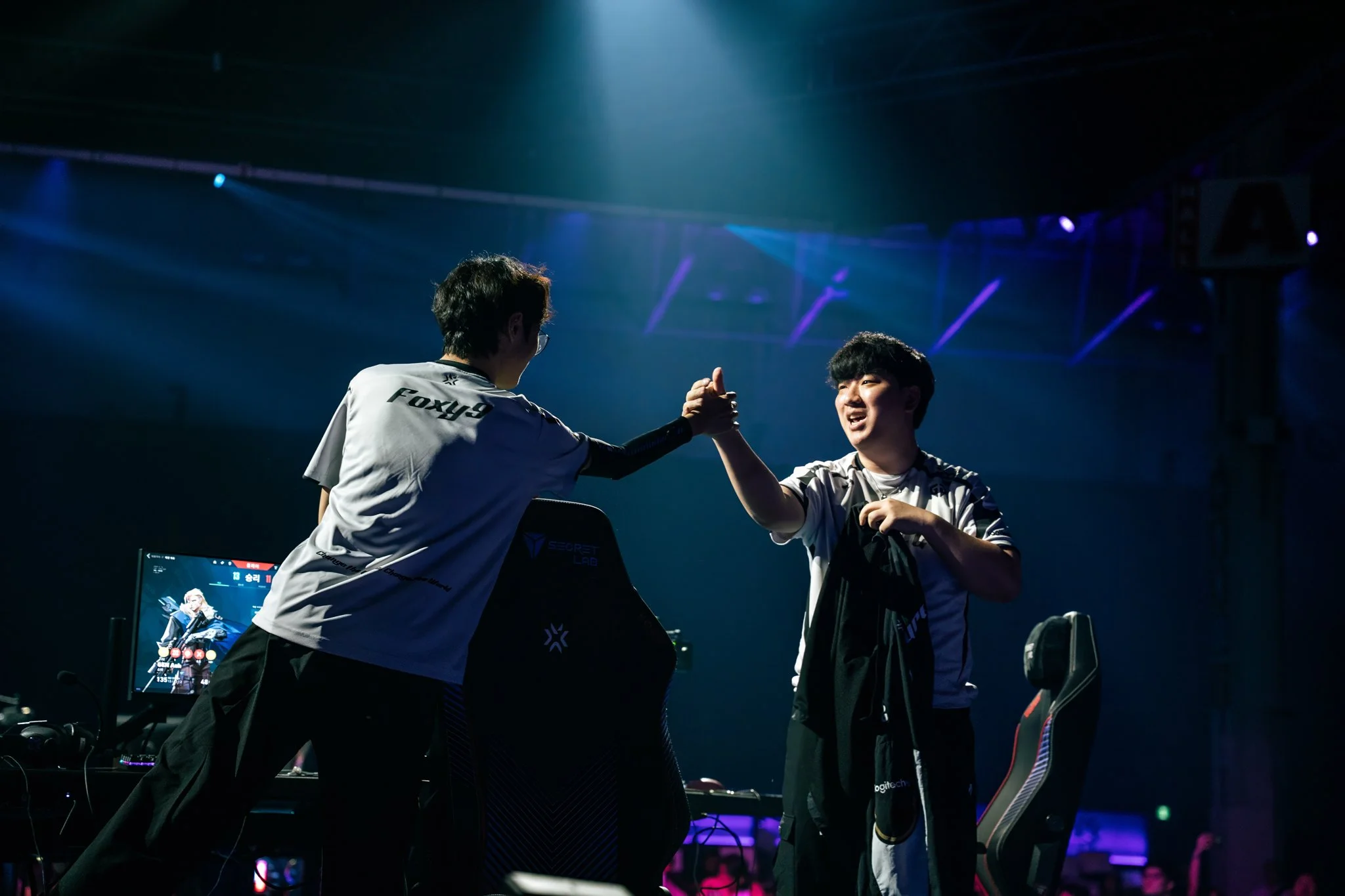 Two esports players in team jerseys exchanging a handshake on stage at an esports event, with gaming equipment and monitors in the background.