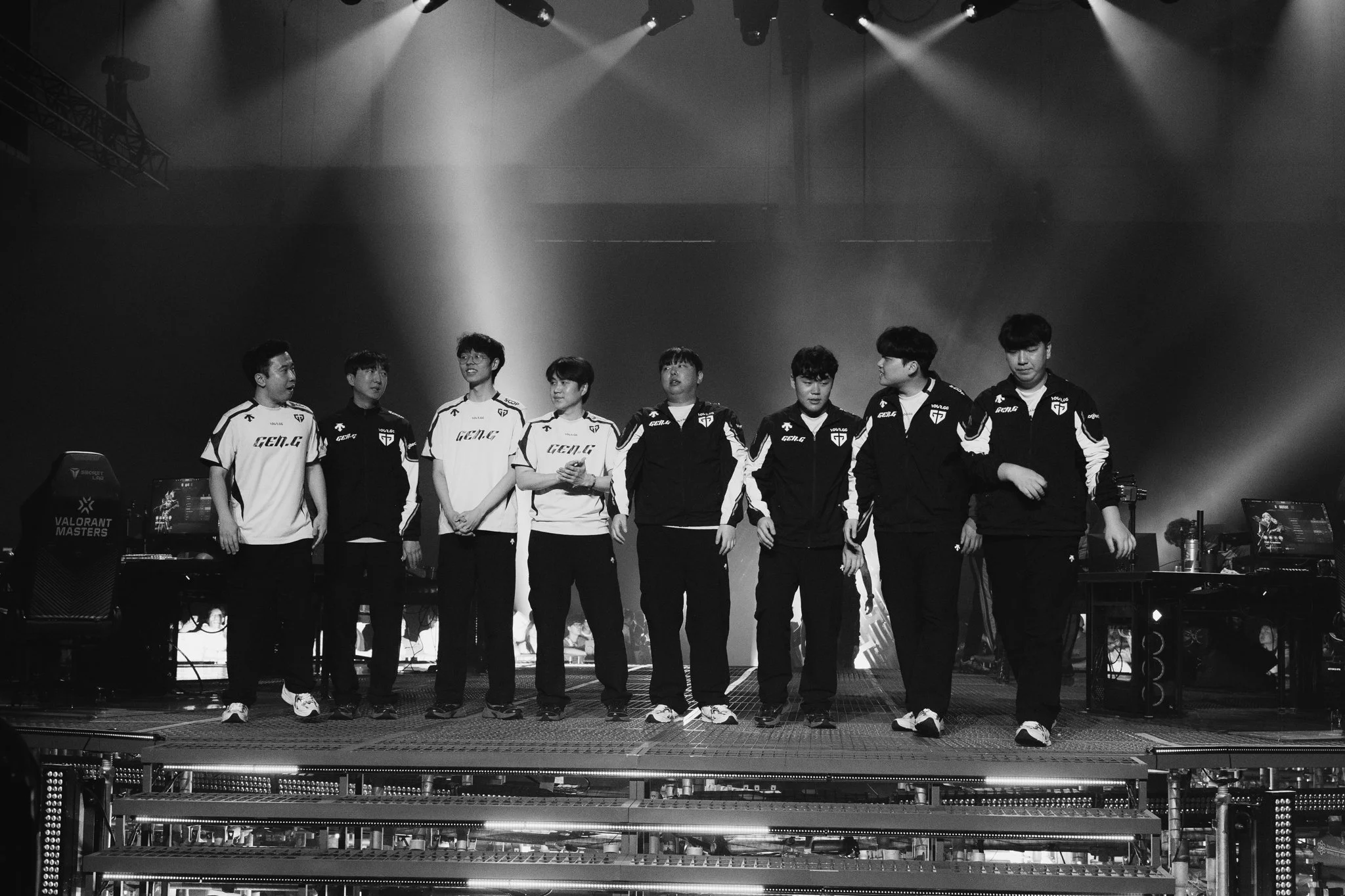 A team of esports players standing on stage at a gaming event, facing the audience, with some looking at each other and some at the crowd, illuminated by stage lights, with gaming equipment and monitors nearby.