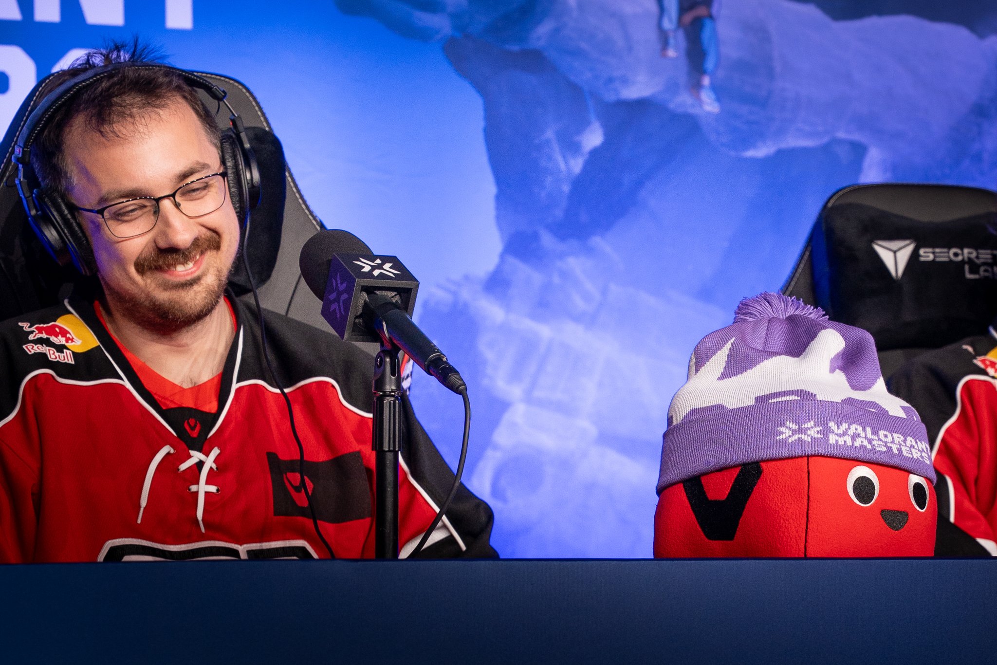 A man in a red esports jersey with a beard, glasses, and headphones is smiling at a microphone. Next to him is a plush toy with a hat, both representing Valorant Masters.