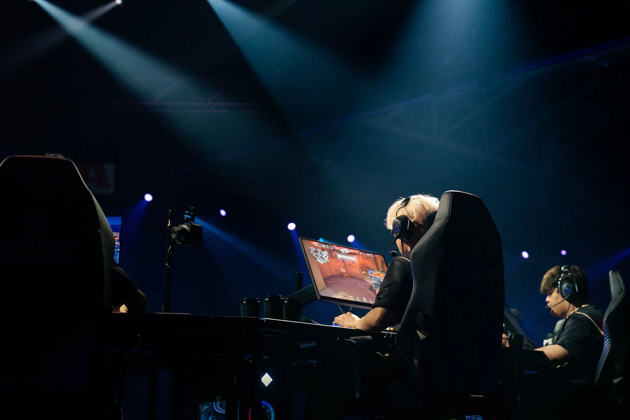 Esports players competing in a professional gaming tournament, wearing headsets and using gaming monitors, illuminated by stage lighting.