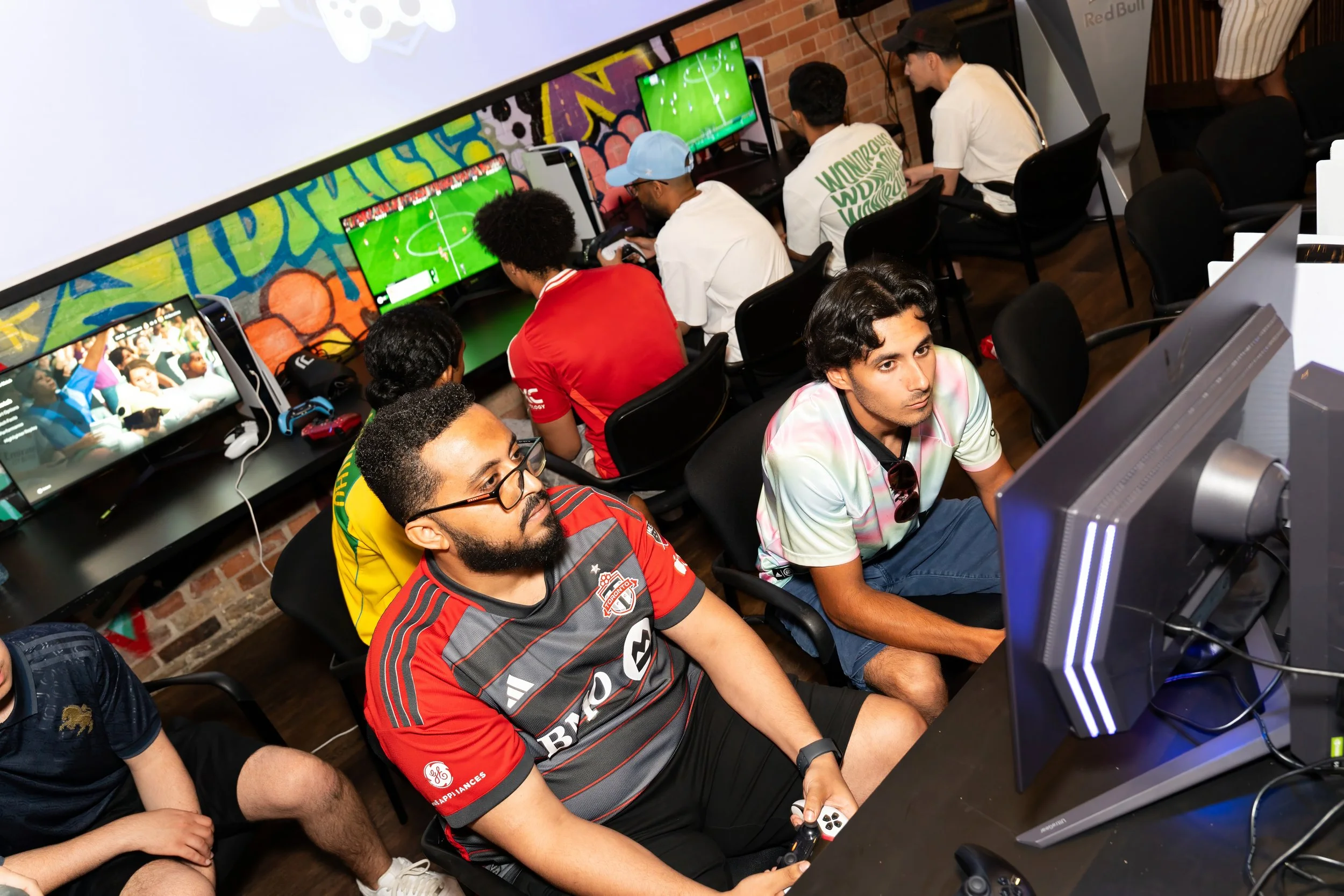 Group of young people playing video games on multiple screens in an arcade or gaming lounge with brick walls and colorful graffiti.