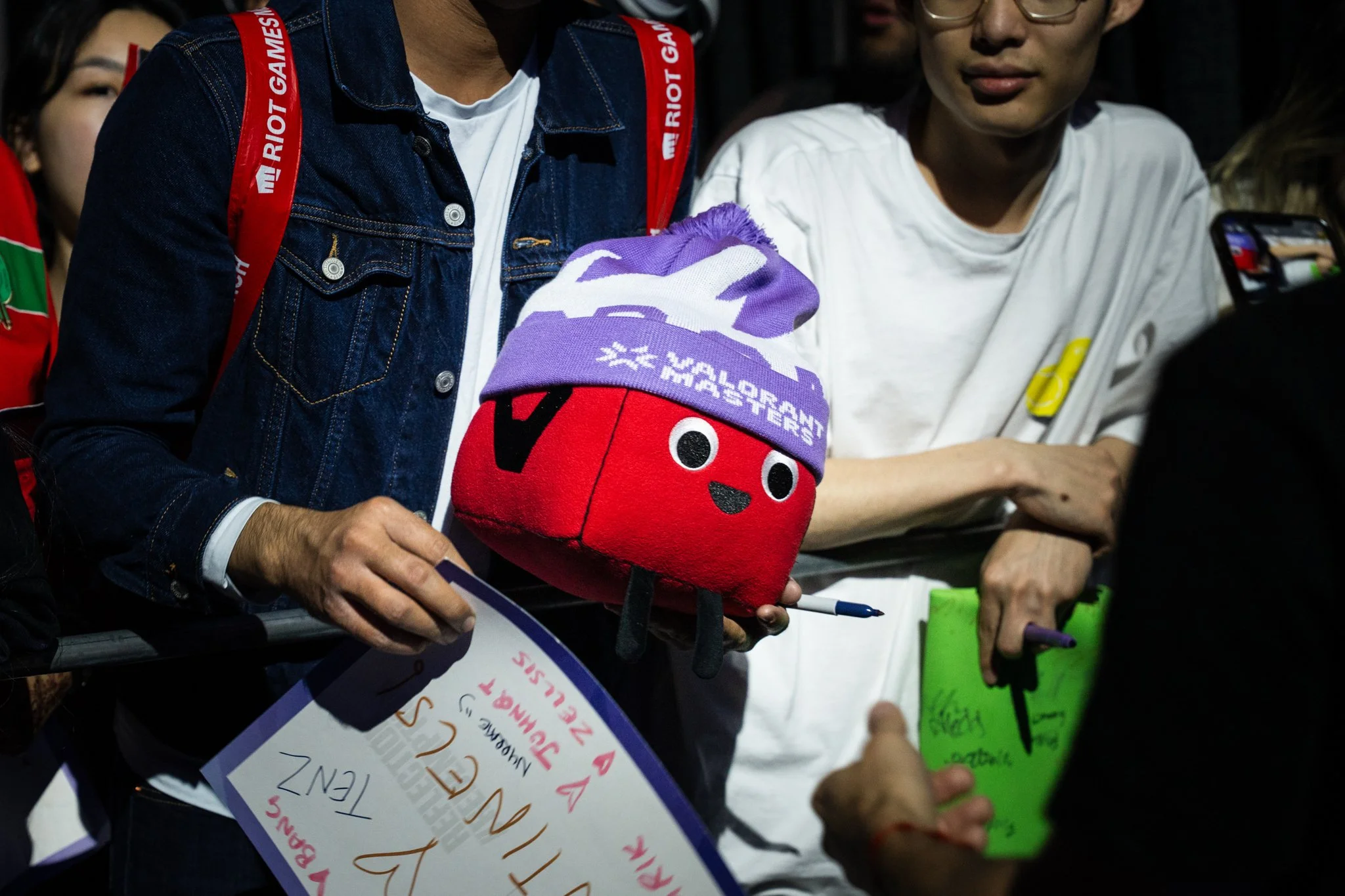 A person holding a plush toy of a red character wearing a purple hat with white writing, surrounded by fans at a public event.