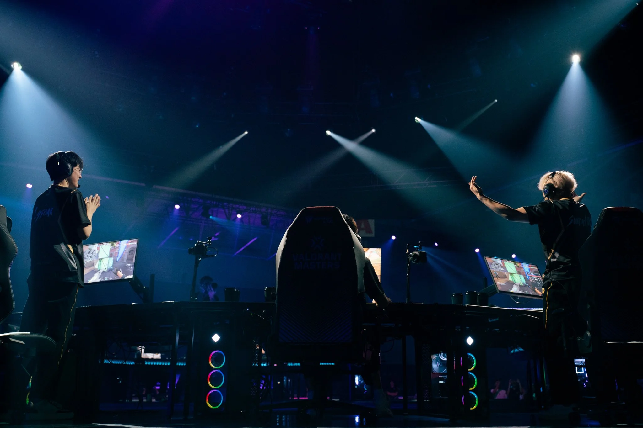 E-sports players in a dimly lit arena with bright spotlights, playing a game at a tournament with screens displaying game footage.