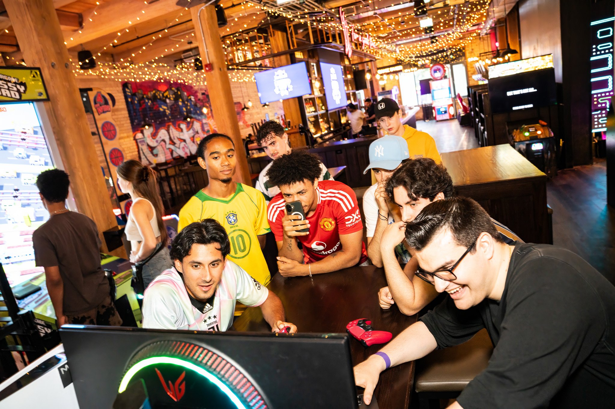 Group of young people gathered around a gaming computer in a lively, decorated arcade or bar, with some watching and others playing video games.