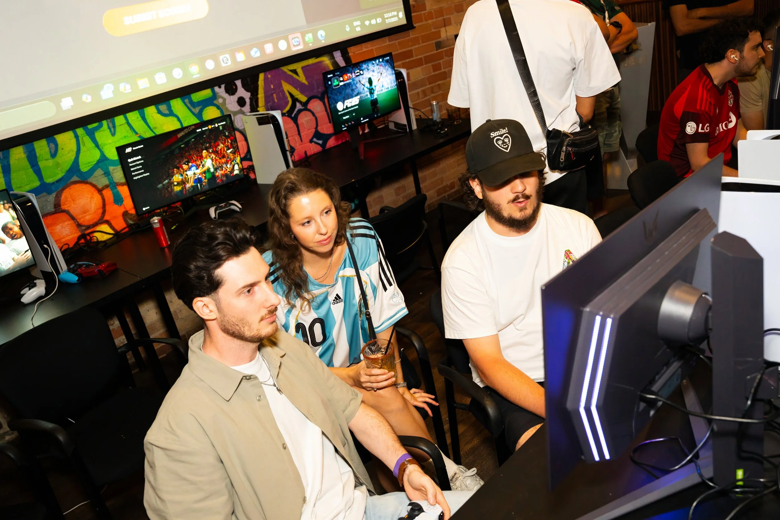 People playing video games at a gaming event, sitting in front of monitors, with a colorful wall in the background.