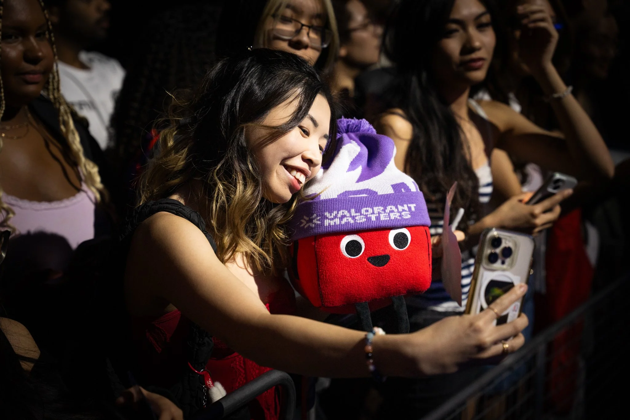 A group of women at a concert or event, with one woman smiling and taking a selfie. She has shoulder-length brown hair with blonde highlights and is wearing a sleeveless top. A woman in the center has a plush purse resembling a red square with a purp